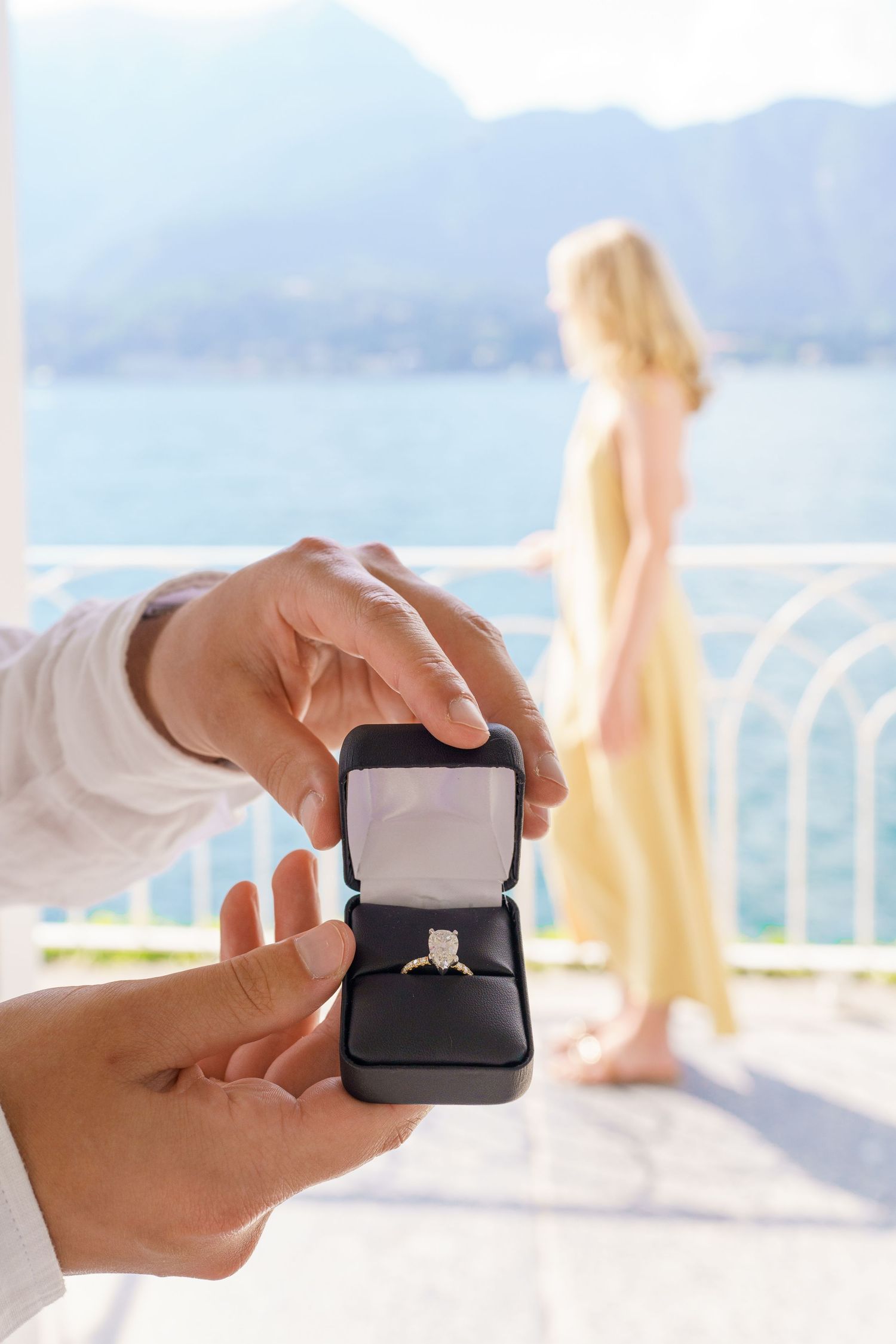 Man holding open ring box with diamond engagement ring, woman in yellow dress standing on scenic lakeside balcony.