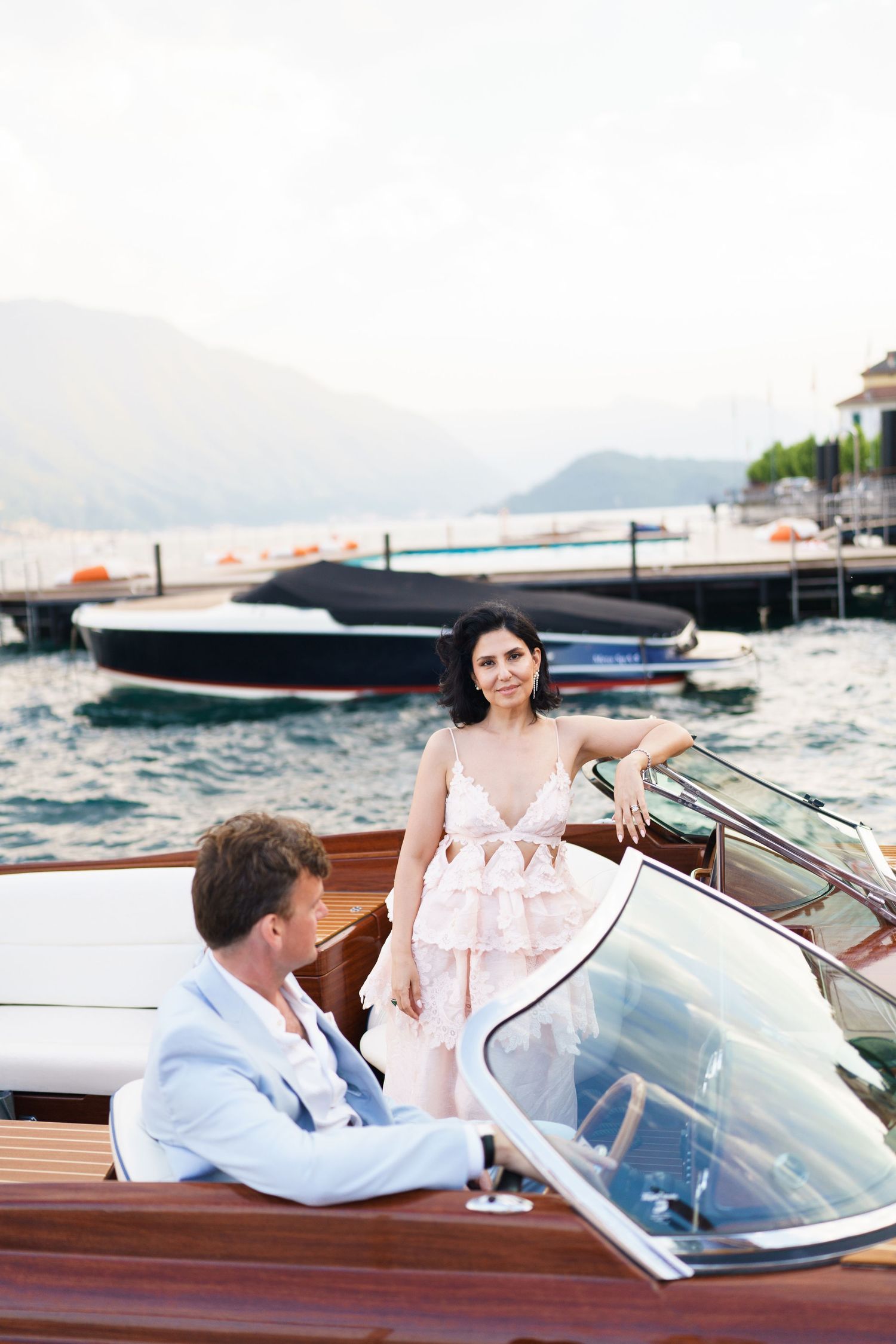 Elegant couple on a luxury wooden speedboat with marina and mountains in background on a calm day.
