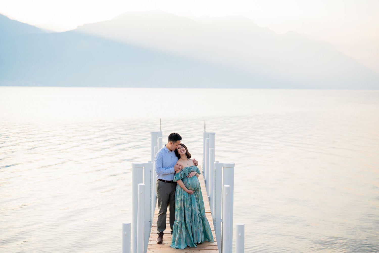 Couple embracing on a white dock over a calm lake with misty mountains, woman in teal gown, romantic outdoor portrait.
