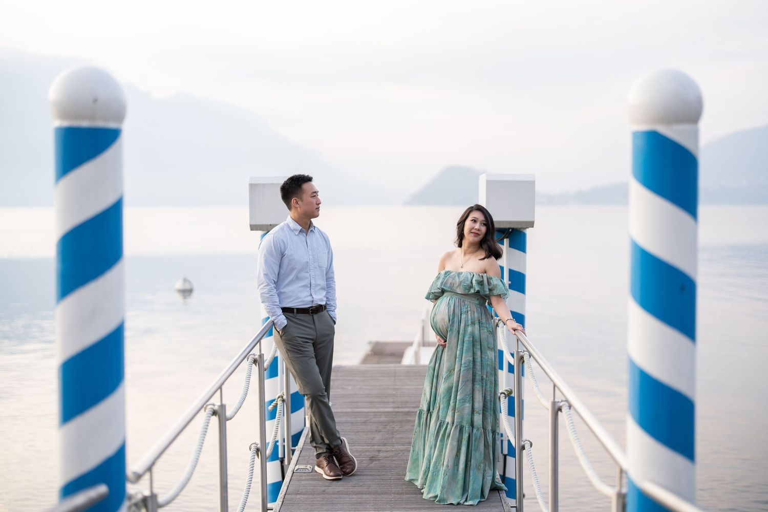 Couple standing on a waterfront dock with blue and white striped poles, overlooking a misty lake and mountains.
