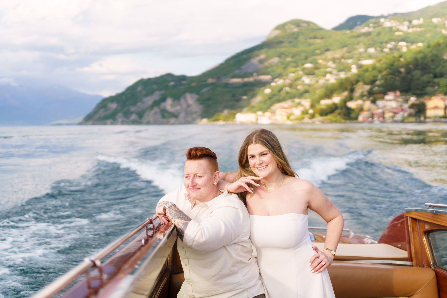 Couple smiling on a wooden speedboat on Lake Como, Italy, with lush green hills and a village in the background.