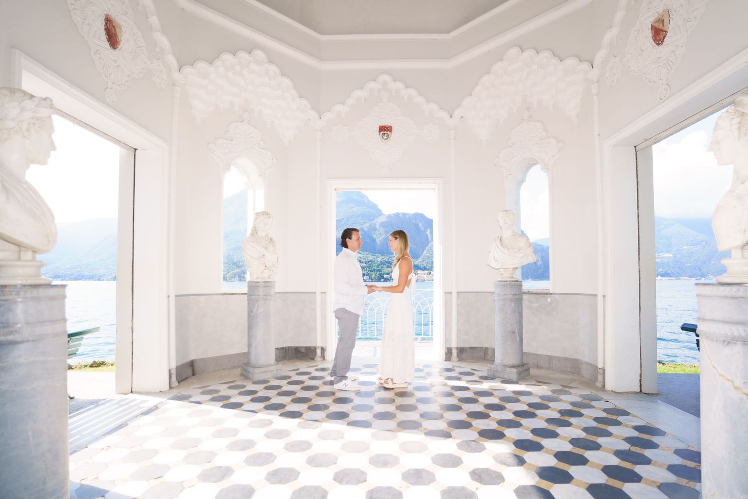 A couple embraces in a white gazebo with checkerboard floor overlooking a scenic lake view.