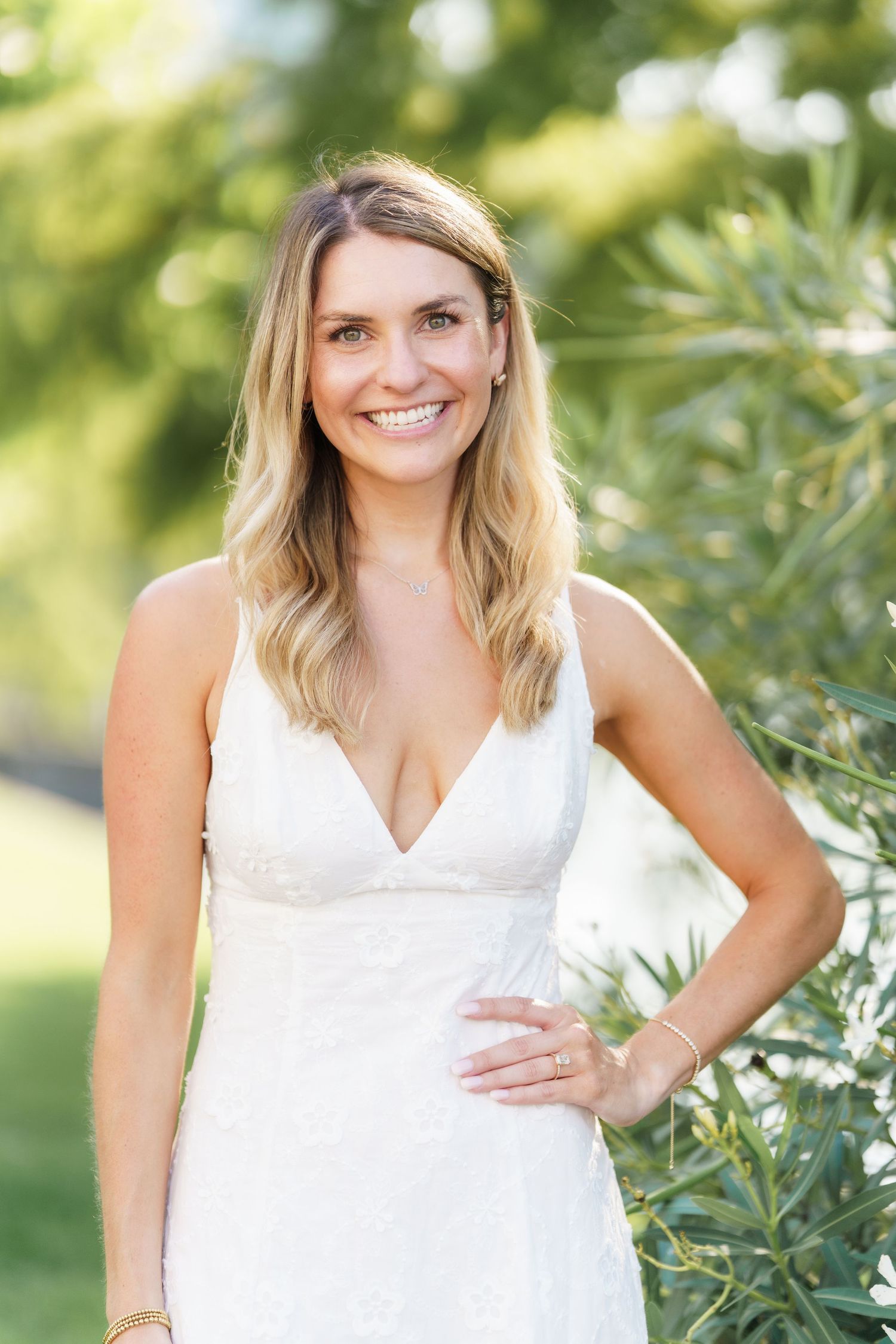 A person wearing a white sleeveless top poses outdoors in a natural setting with greenery in the background.