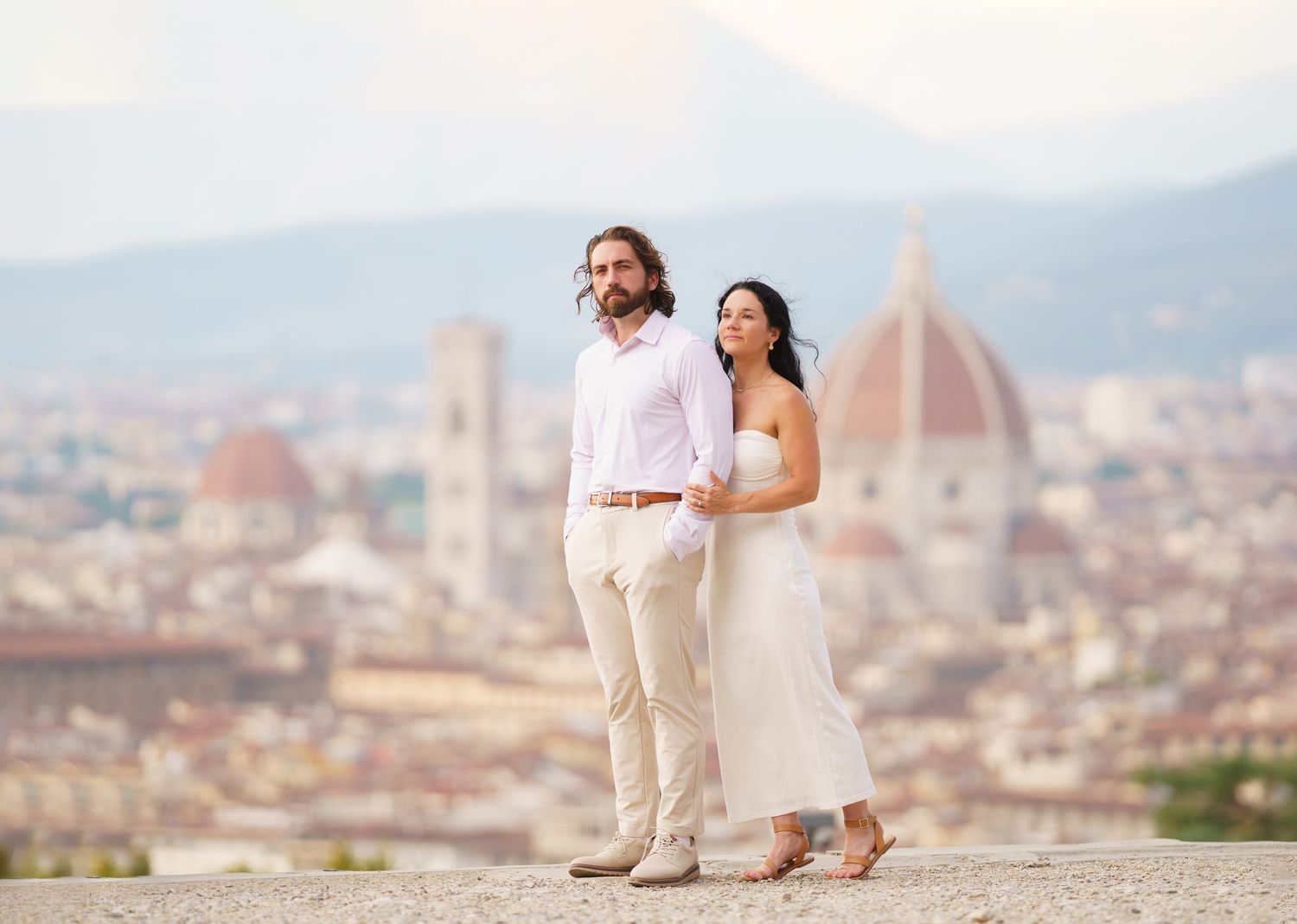Couple sits on stone wall overlooking Florence cityscape with Duomo cathedral visible in background.