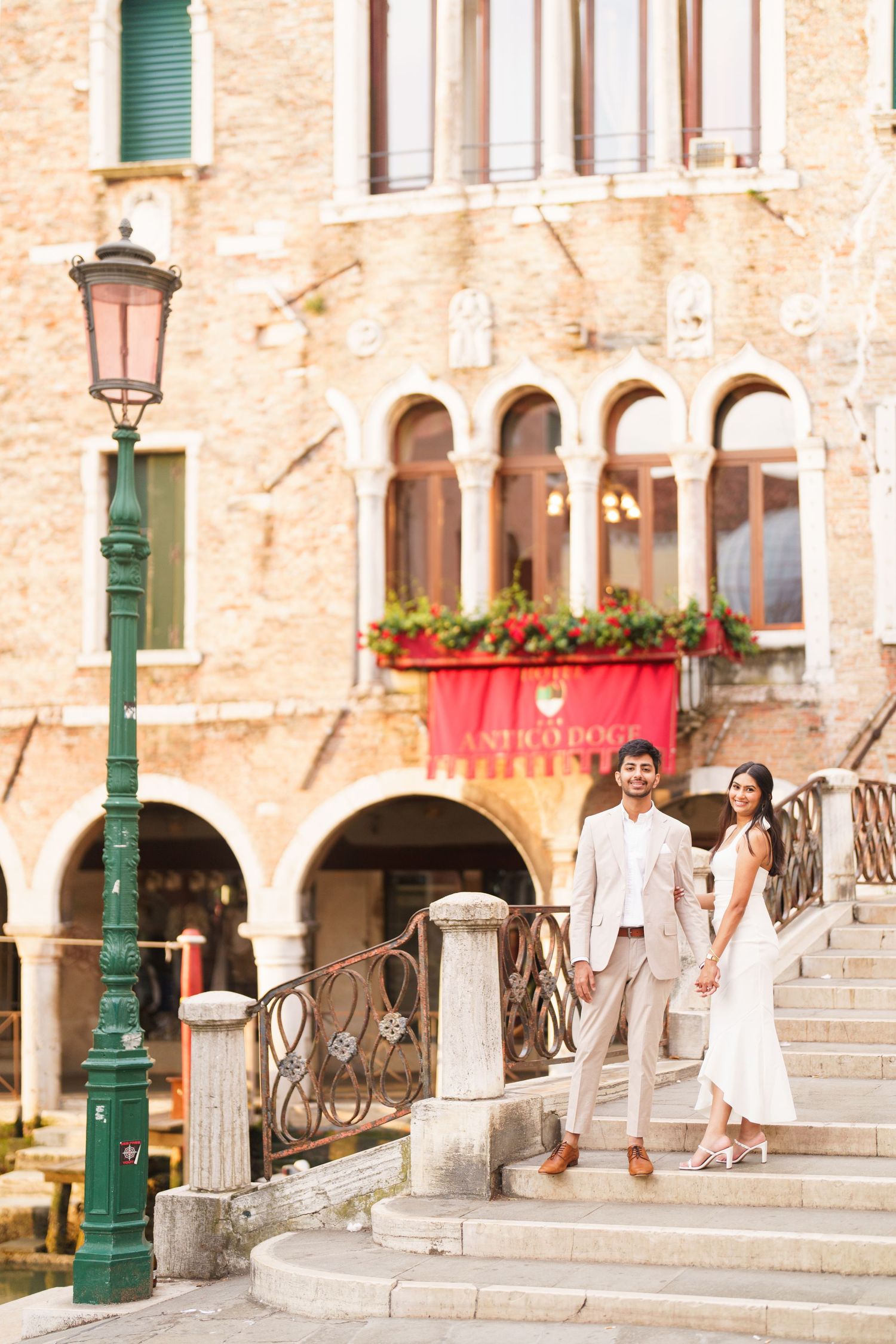 A couple in white outfits walks together on stone steps near historic Mediterranean architecture with green shutters.