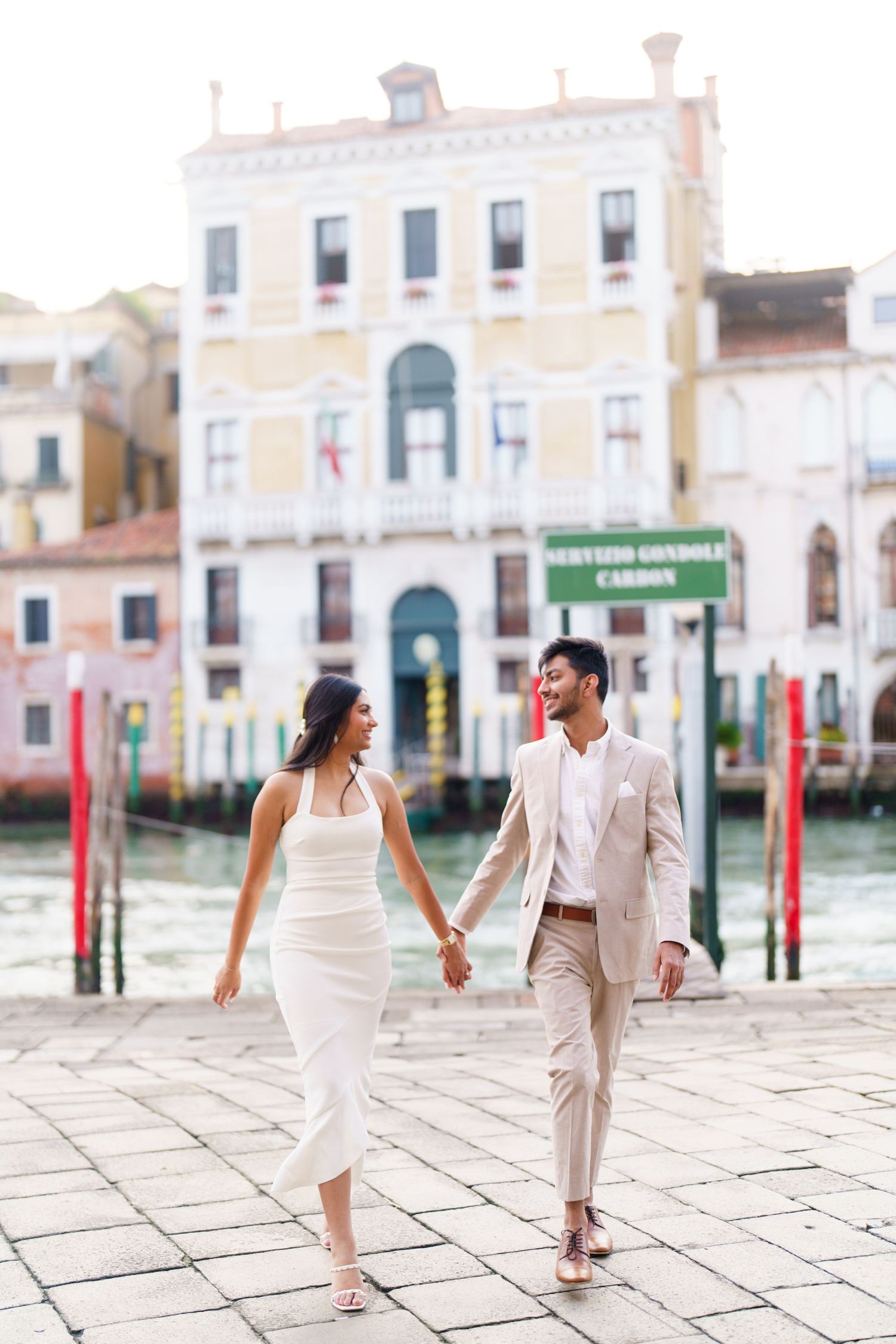 A couple in elegant white and beige outfits walk hand in hand along Venice's historic waterfront.