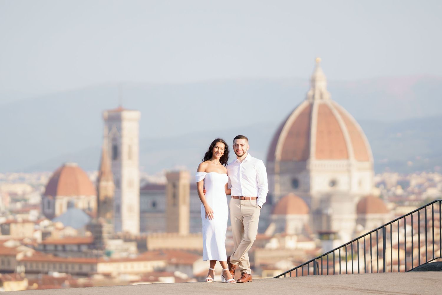 A couple in white outfits poses on a terrace overlooking Florence's iconic Duomo cathedral and historic skyline.