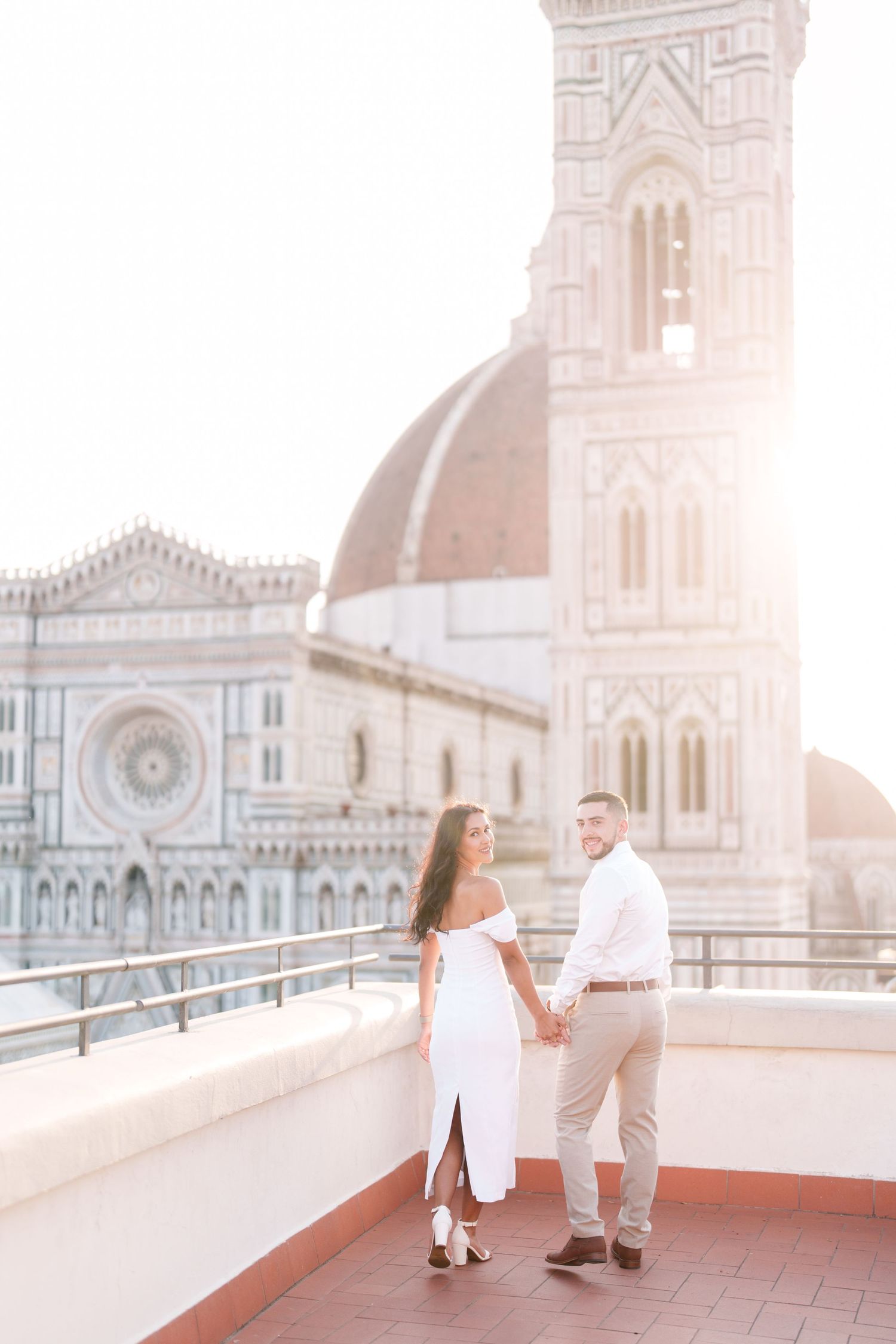 Couple in white outfits holding hands on a rooftop terrace overlooking the Florence Cathedral in Italy during sunset.