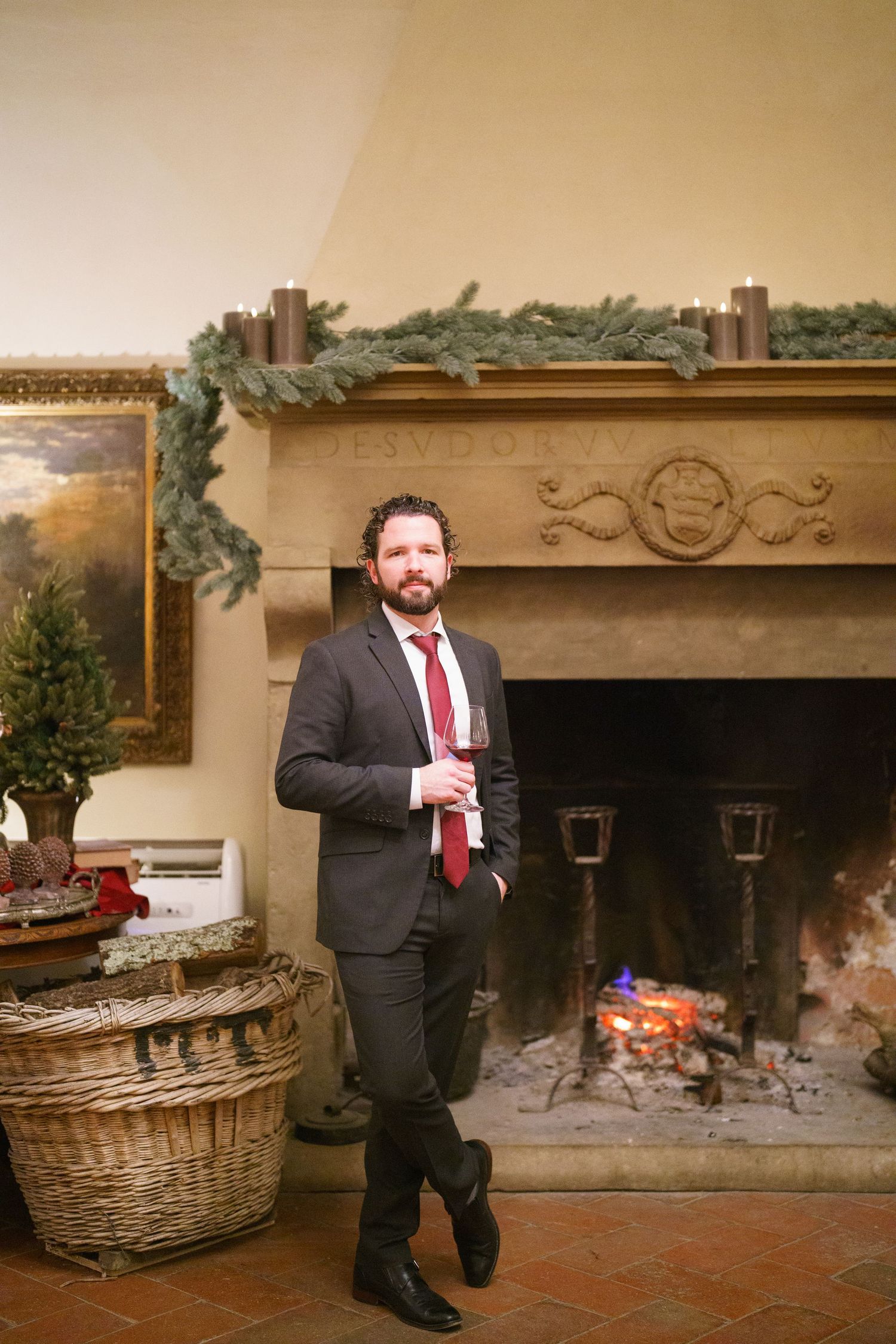 A groom in a black suit and red tie stands by a large stone fireplace decorated for the holidays.