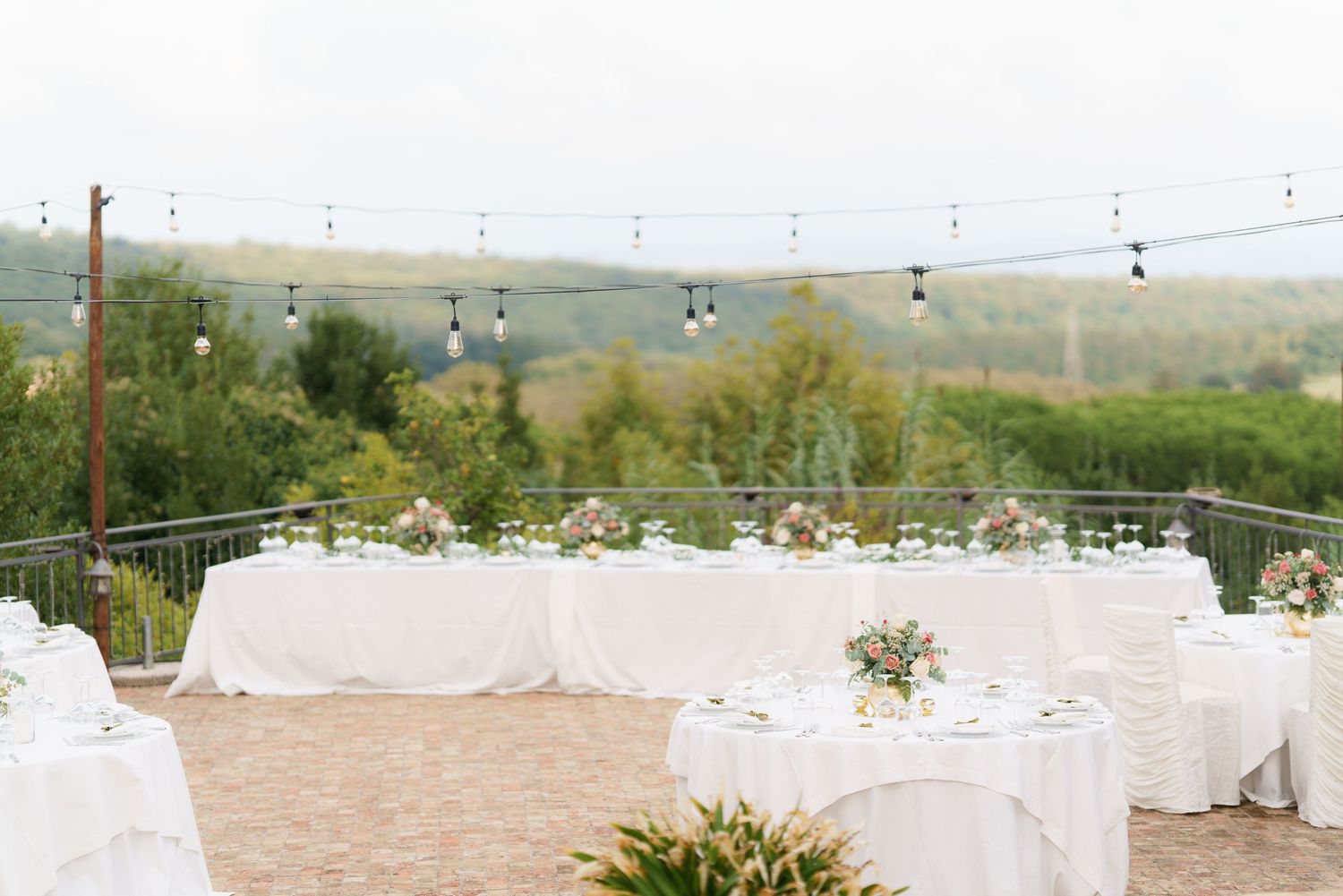 Outdoor wedding reception setup with string lights and white tablecloths against a scenic landscape.