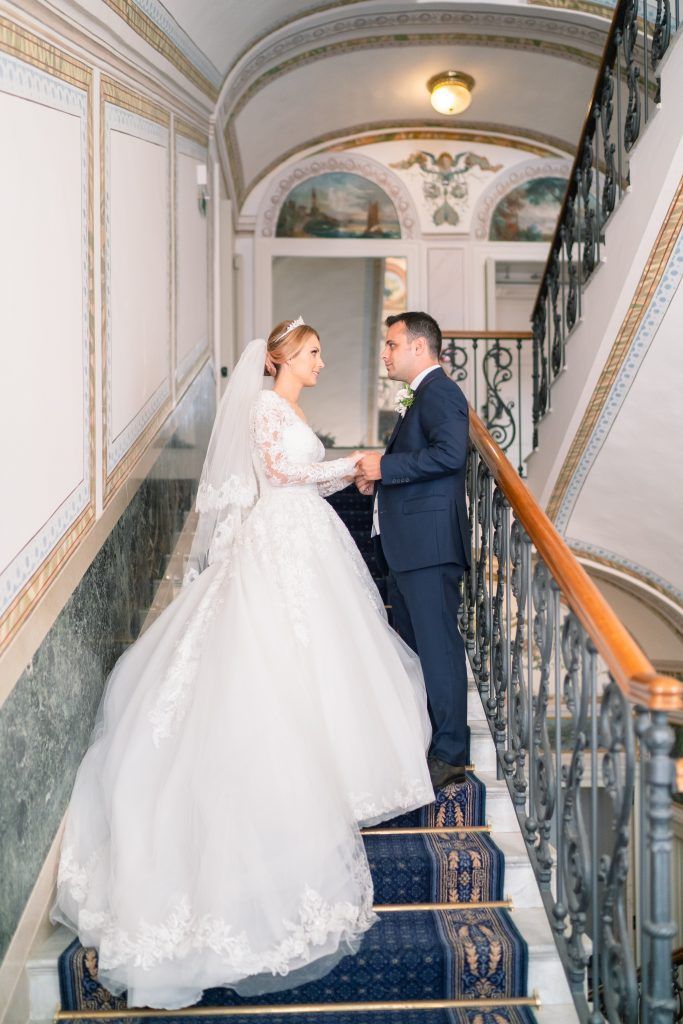 Wedding couple ascending grand marble staircase with ornate railings and arched ceiling.