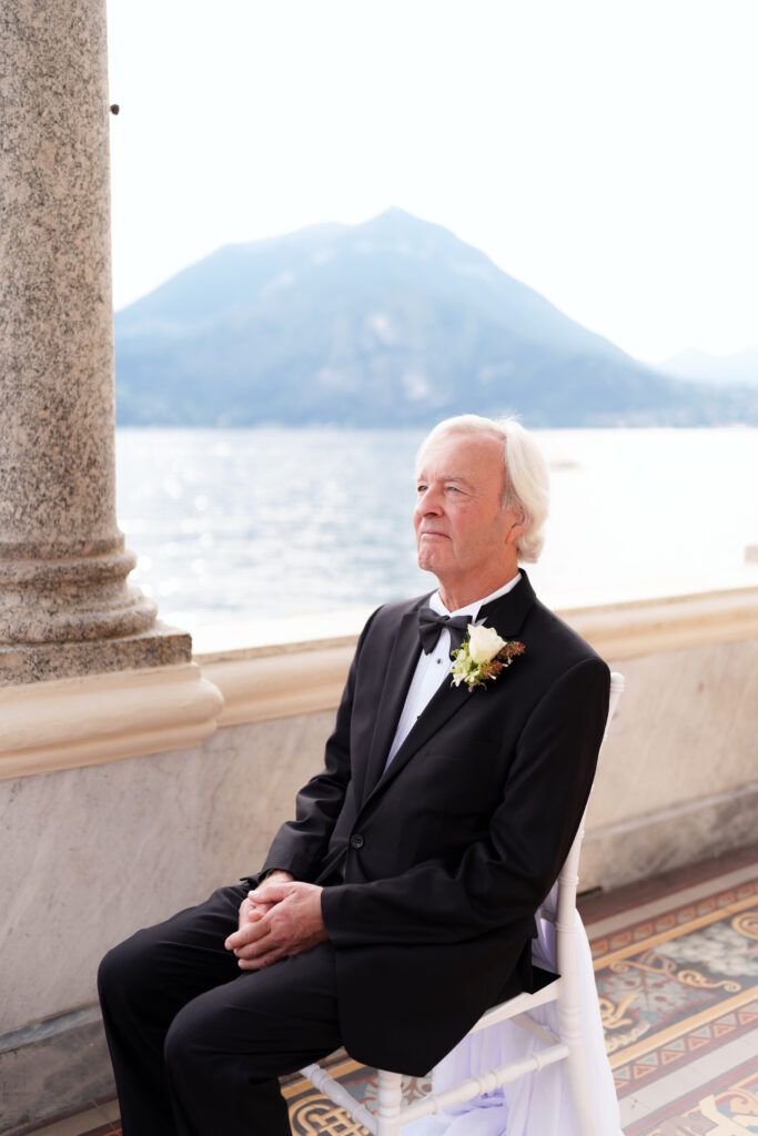 Elderly person in formal black attire sitting on an elegant terrace overlooking mountains and water.
