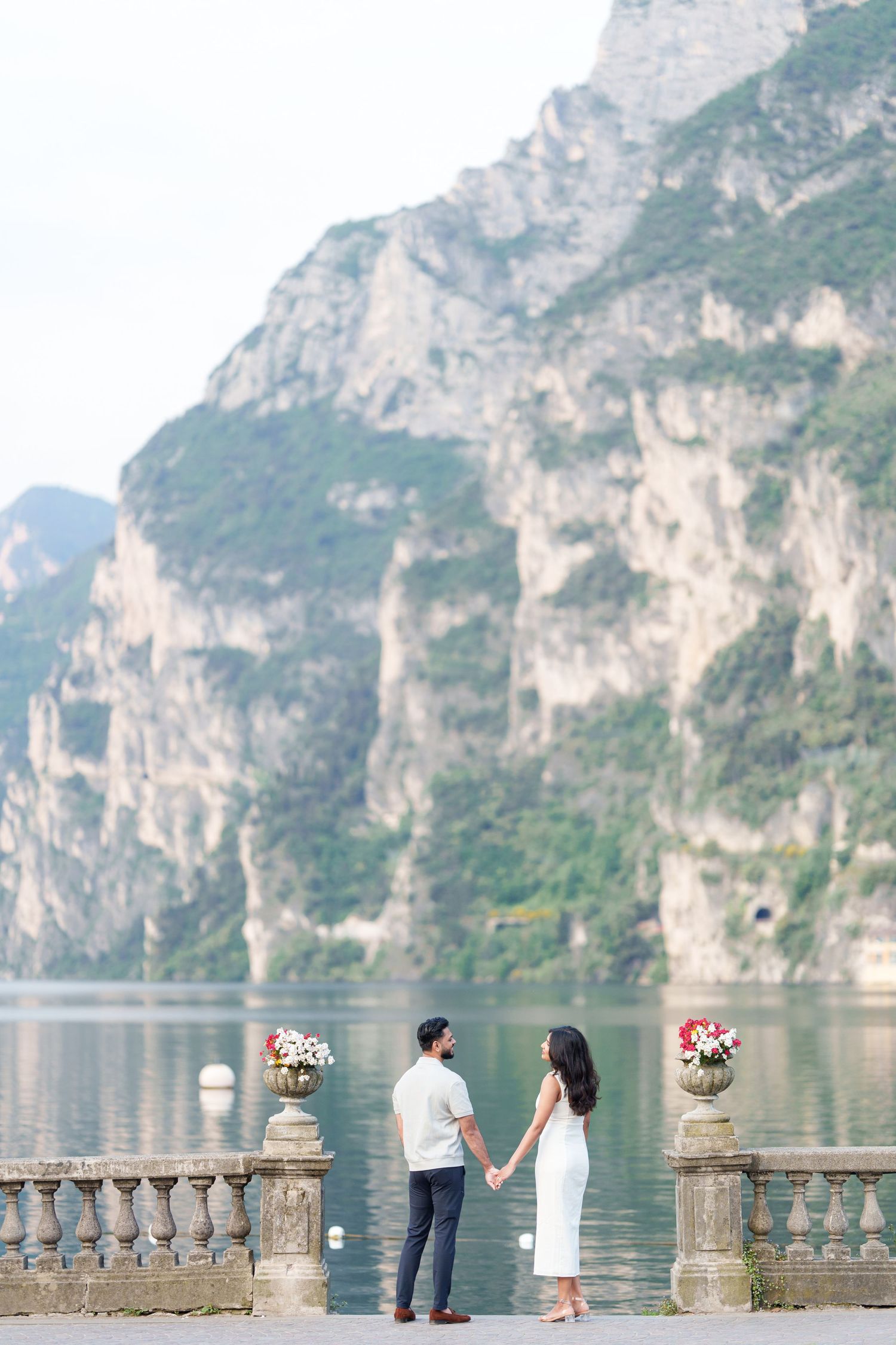 Couple holding hands on stone balcony overlooking scenic Lake Como with dramatic mountain cliffs in background.
