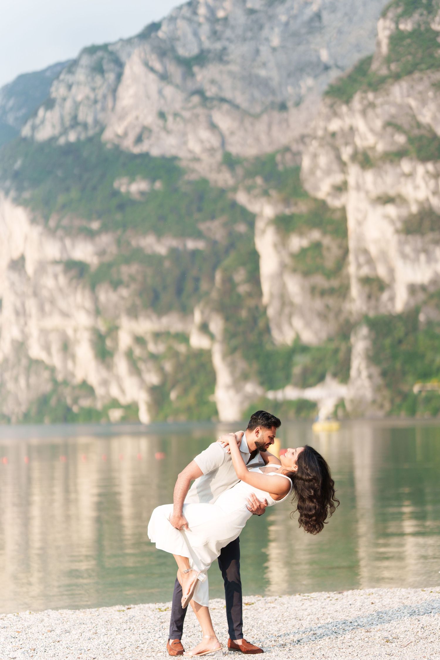 Romantic couple embraces in dramatic dip pose along scenic mountain lake shore with towering cliffs in background.