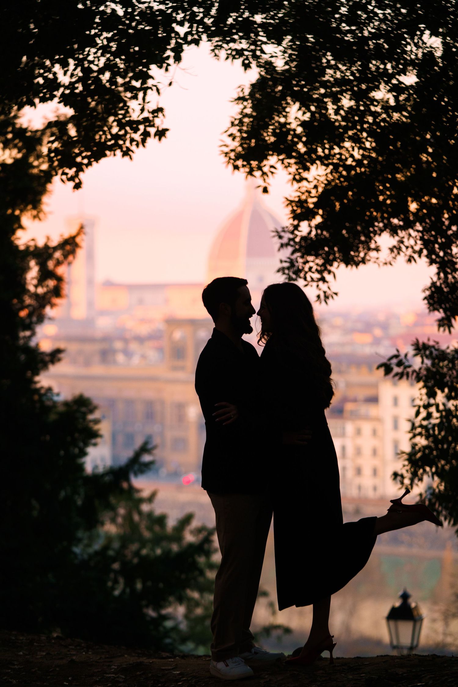 Silhouettes of a romantic couple kissing against a sunset cityscape with tree branches framing the scene.