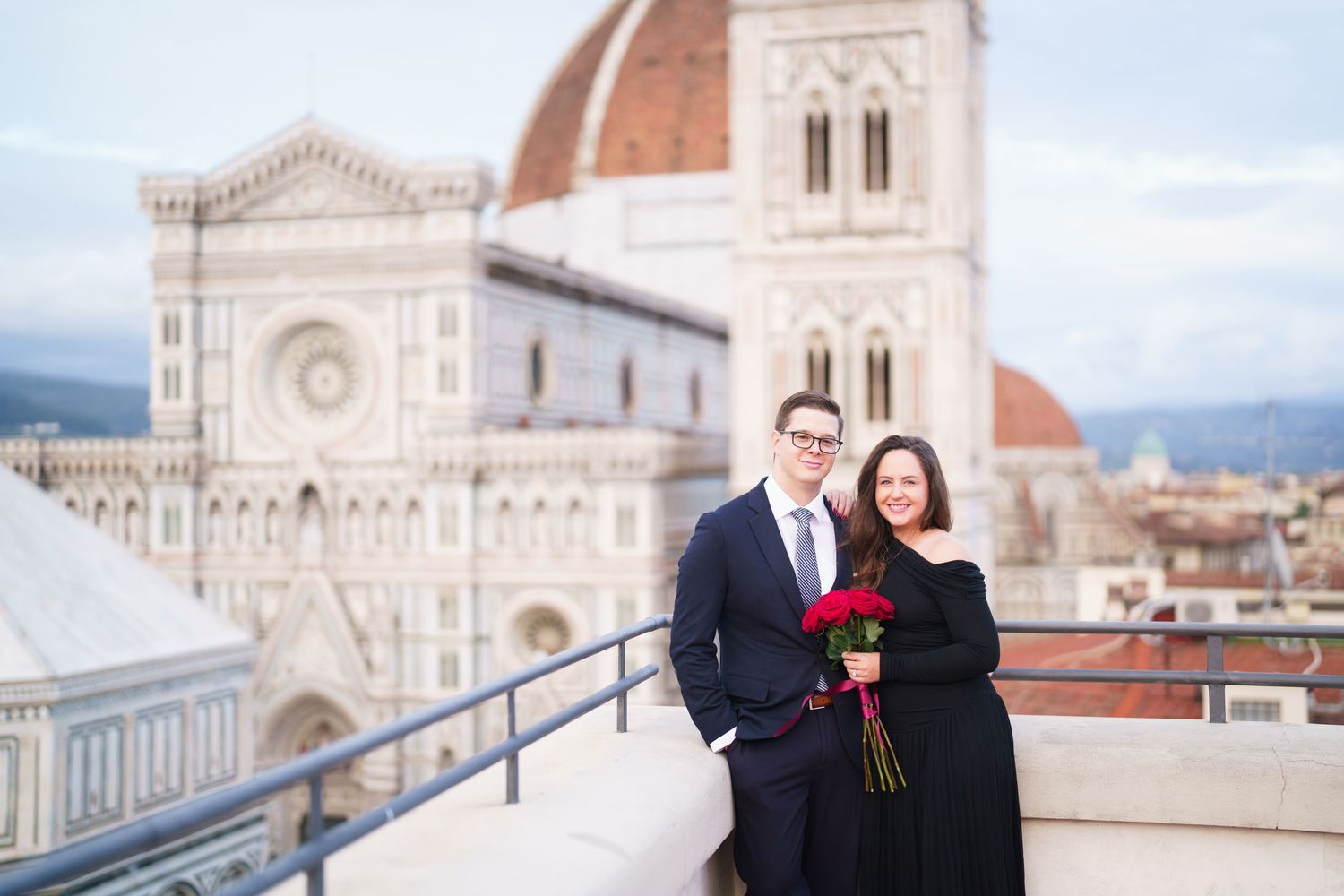 A couple poses on a terrace overlooking the Florence Cathedral and Duomo in Italy on a sunny day.