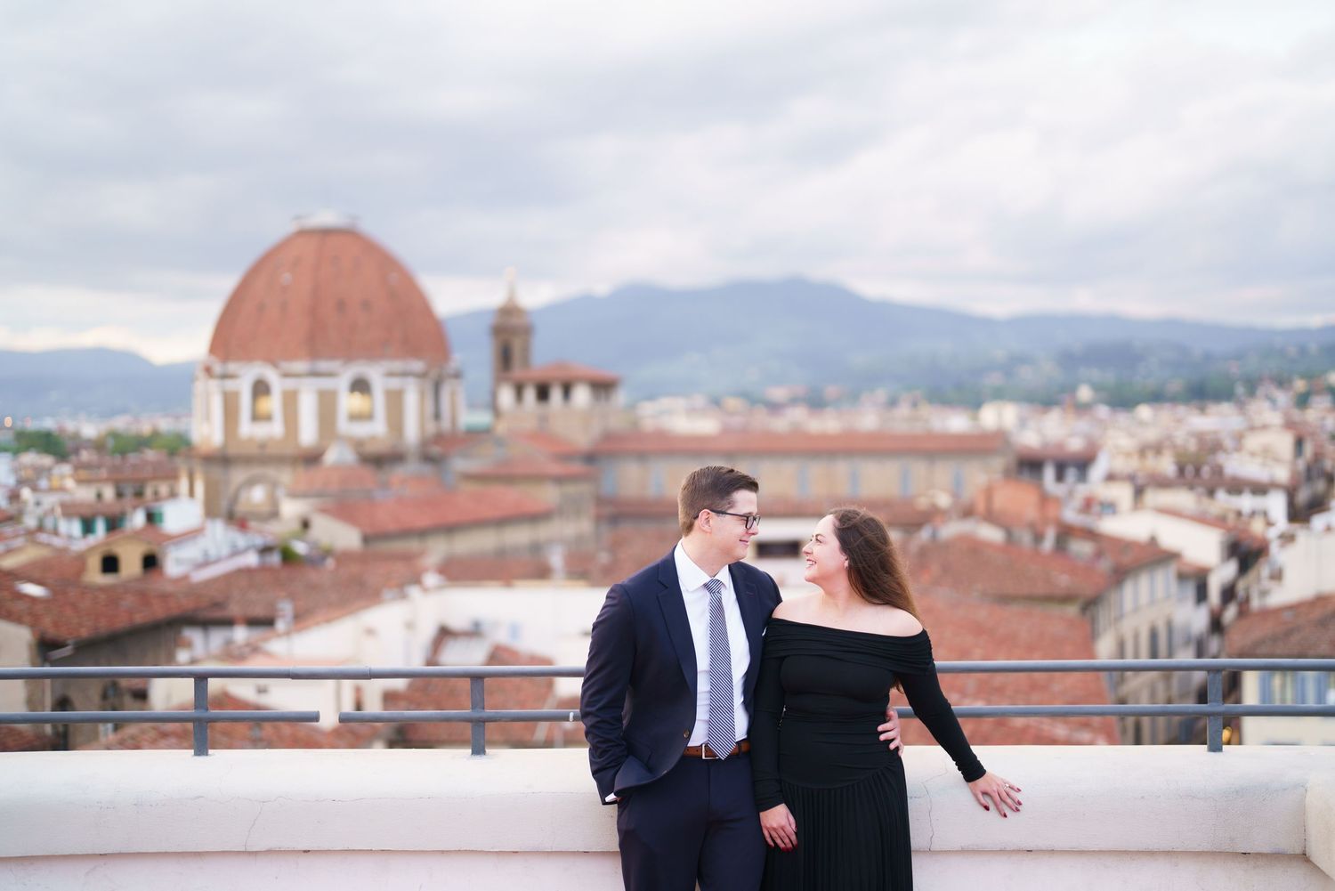 A couple embraces on a rooftop terrace overlooking Florence, Italy with the historic Duomo cathedral in the background.