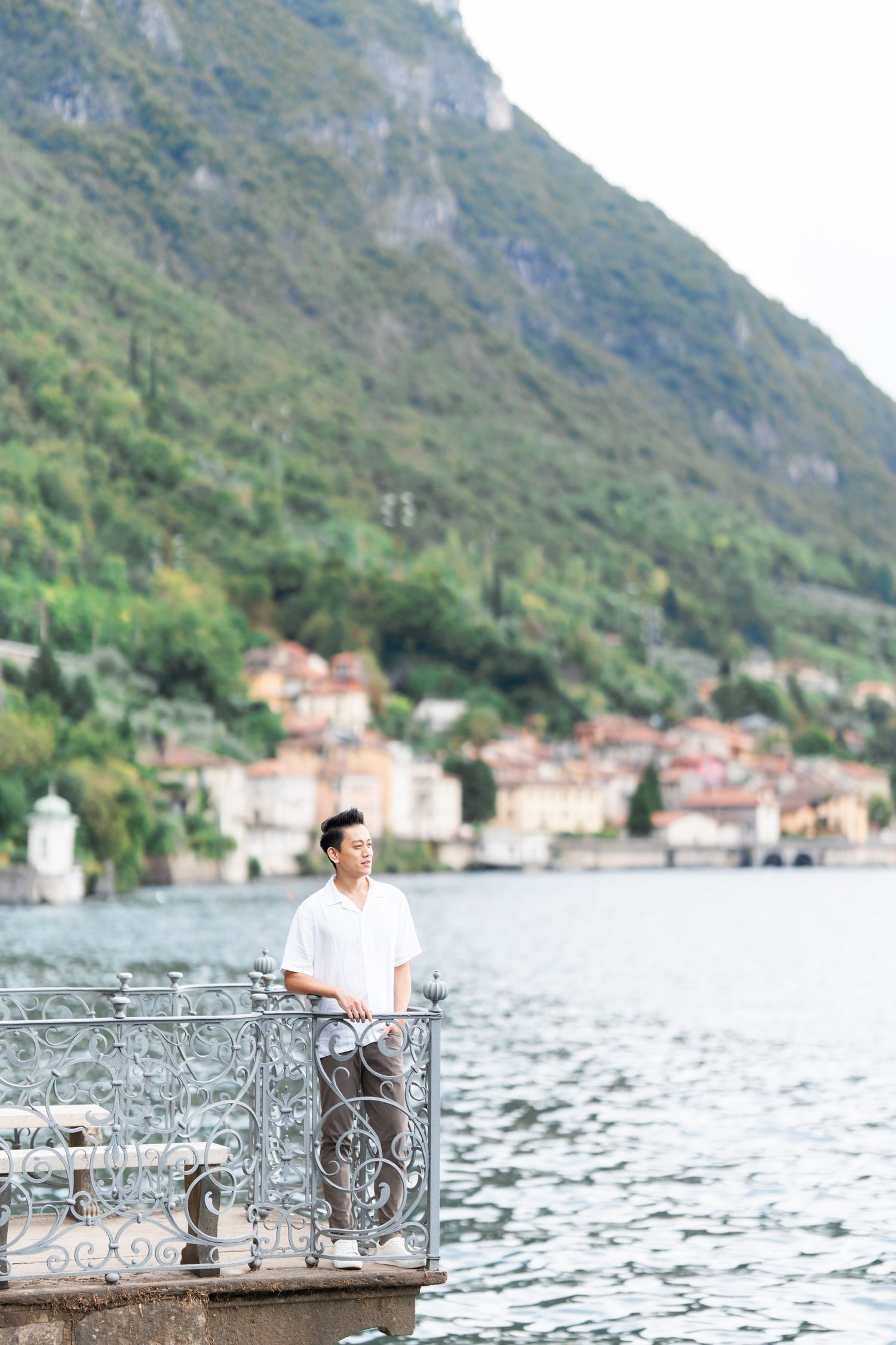 A person sits at a decorative metal table overlooking a scenic lake with mountains and coastal buildings in the background.