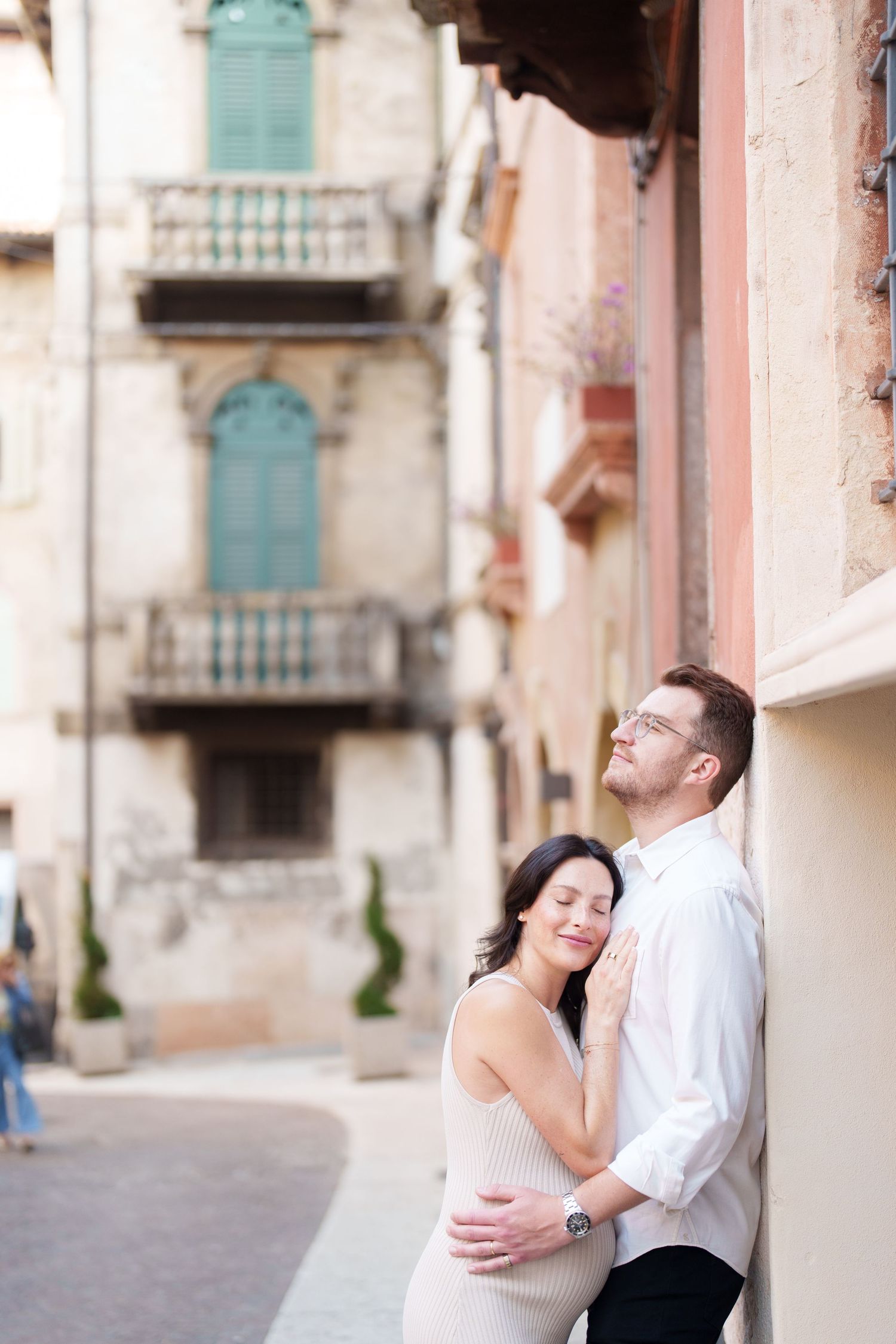 A romantic couple embraces and laughs together against a rustic Mediterranean building with turquoise shutters.