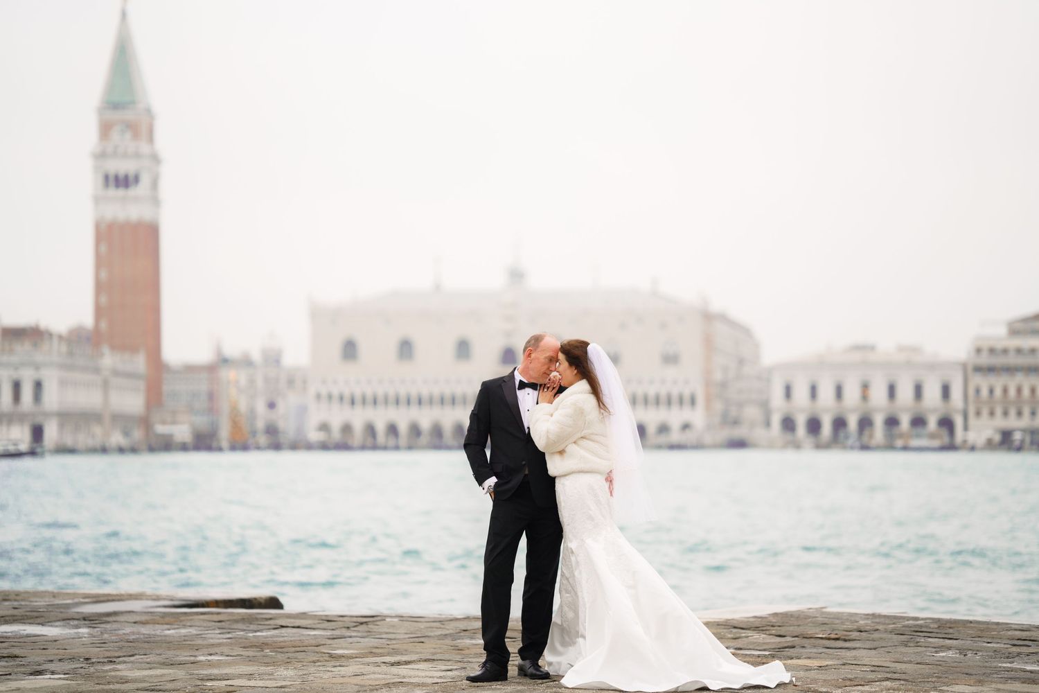 A romantic wedding couple embraces on a pier overlooking Venice's iconic St Mark's Square and Campanile tower.