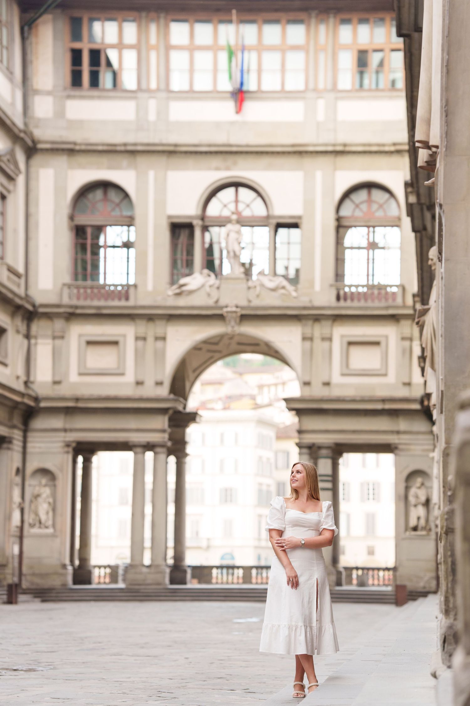 A person in a white dress walks through the elegant arched corridors of the Uffizi Gallery in Florence, Italy.