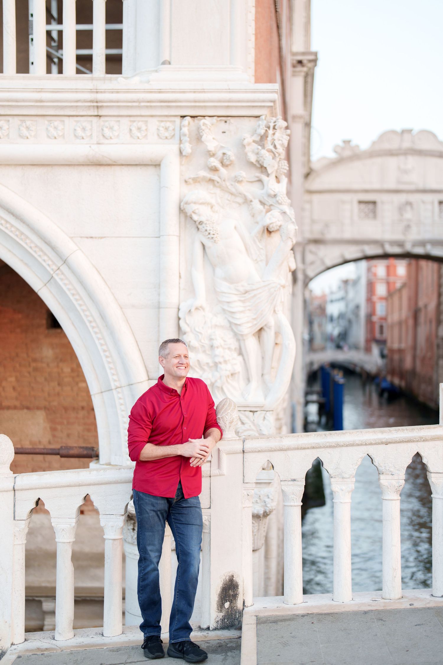 Person in red shirt and jeans poses along ornate white stone balustrade overlooking Venice canal at sunset.
