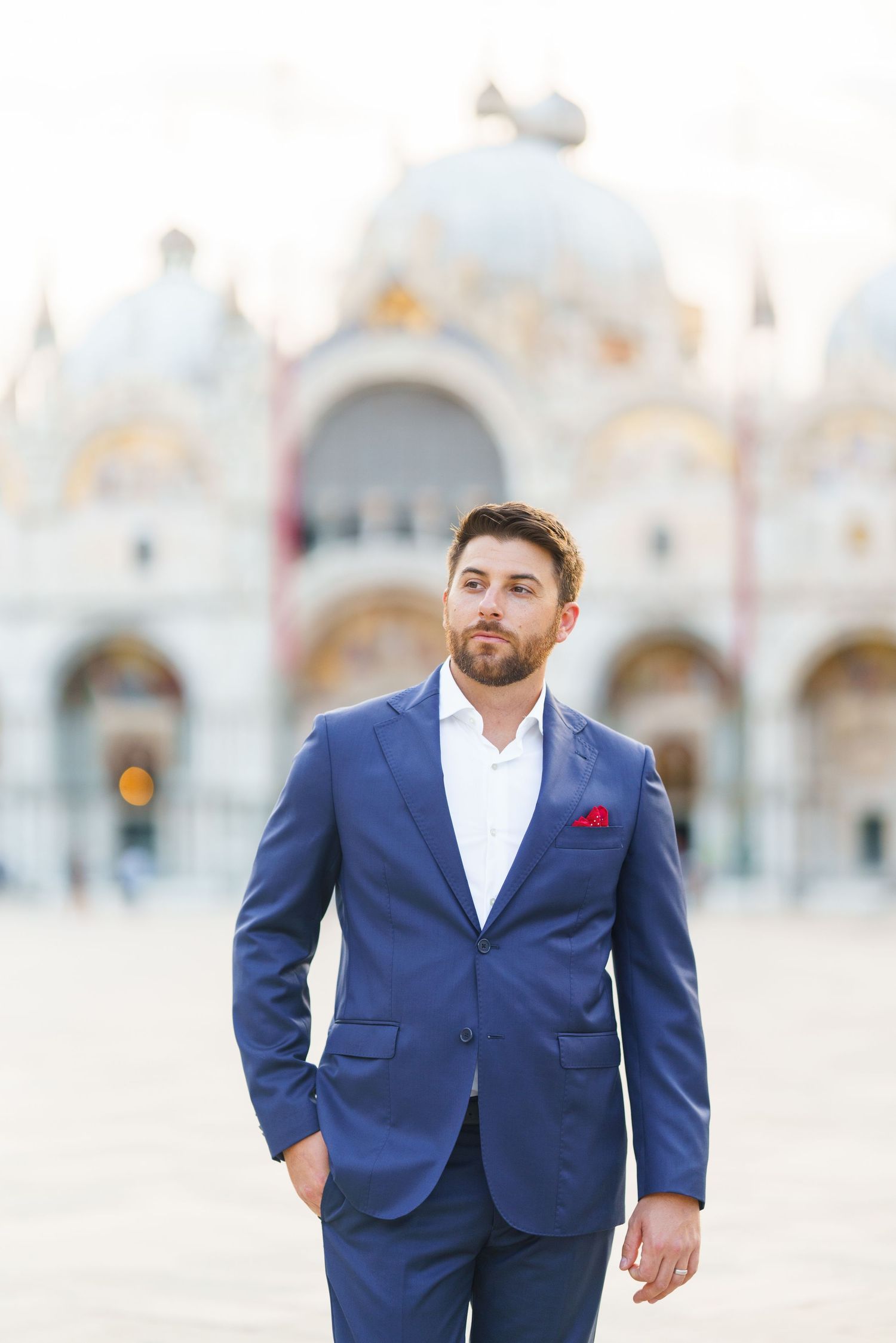 A man in a navy blue suit and white shirt poses outdoors against a blurred architectural background.