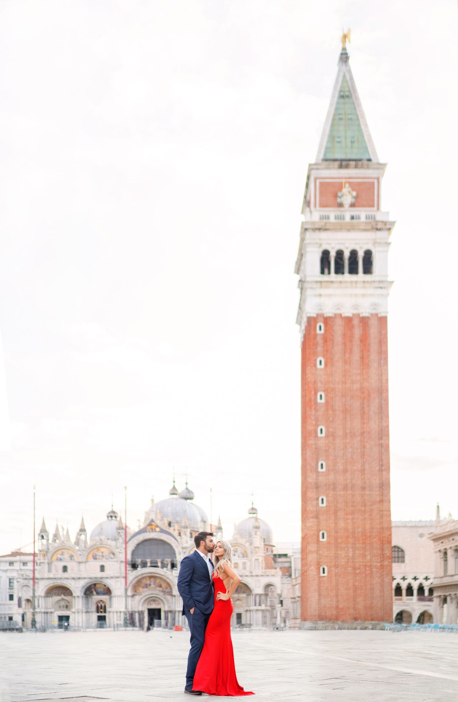 A couple in formal attire shares a romantic moment in Venice's iconic St. Mark's Square with the Campanile tower behind them.