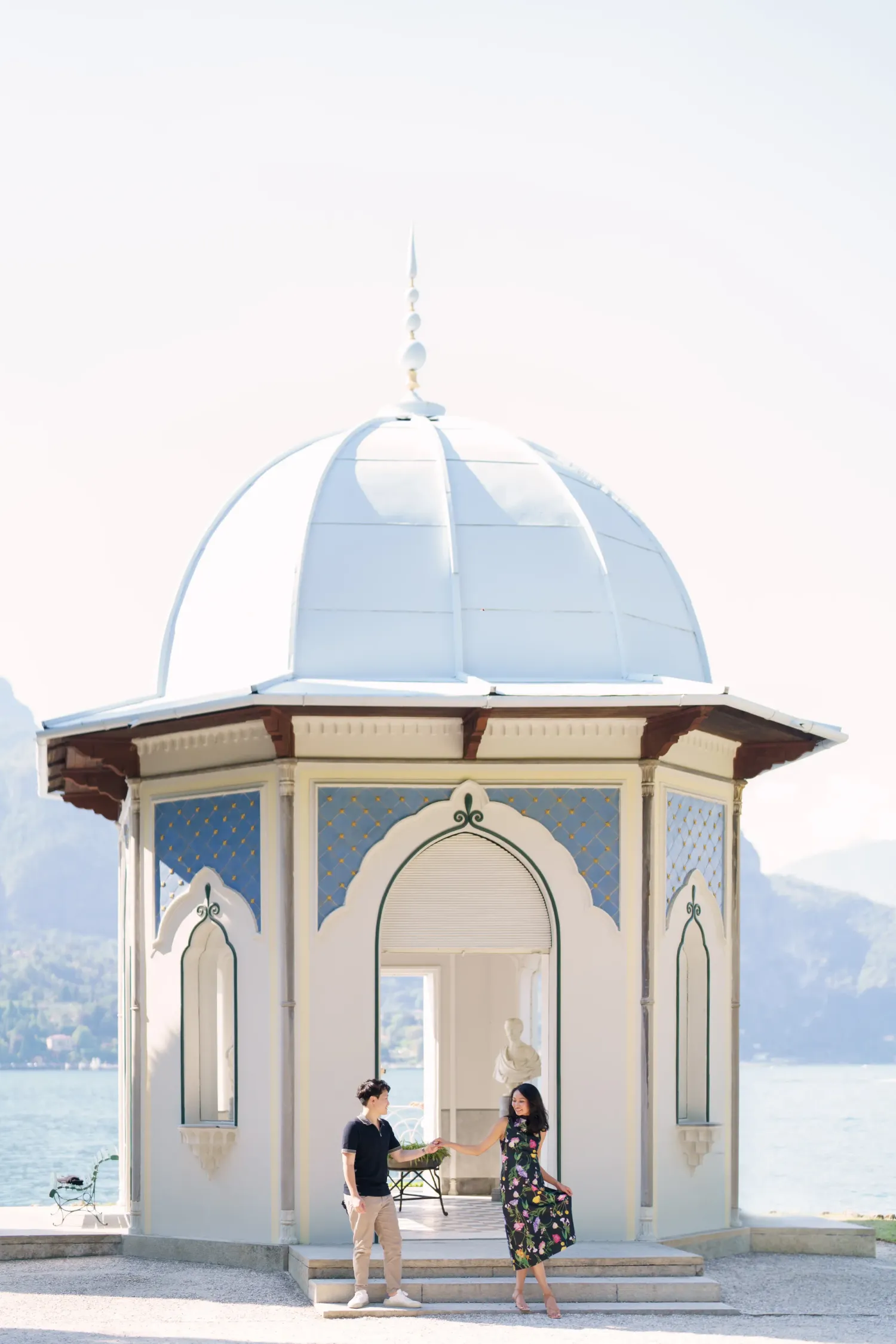Two people stand in a white domed gazebo with ornate arches overlooking a scenic waterfront view.