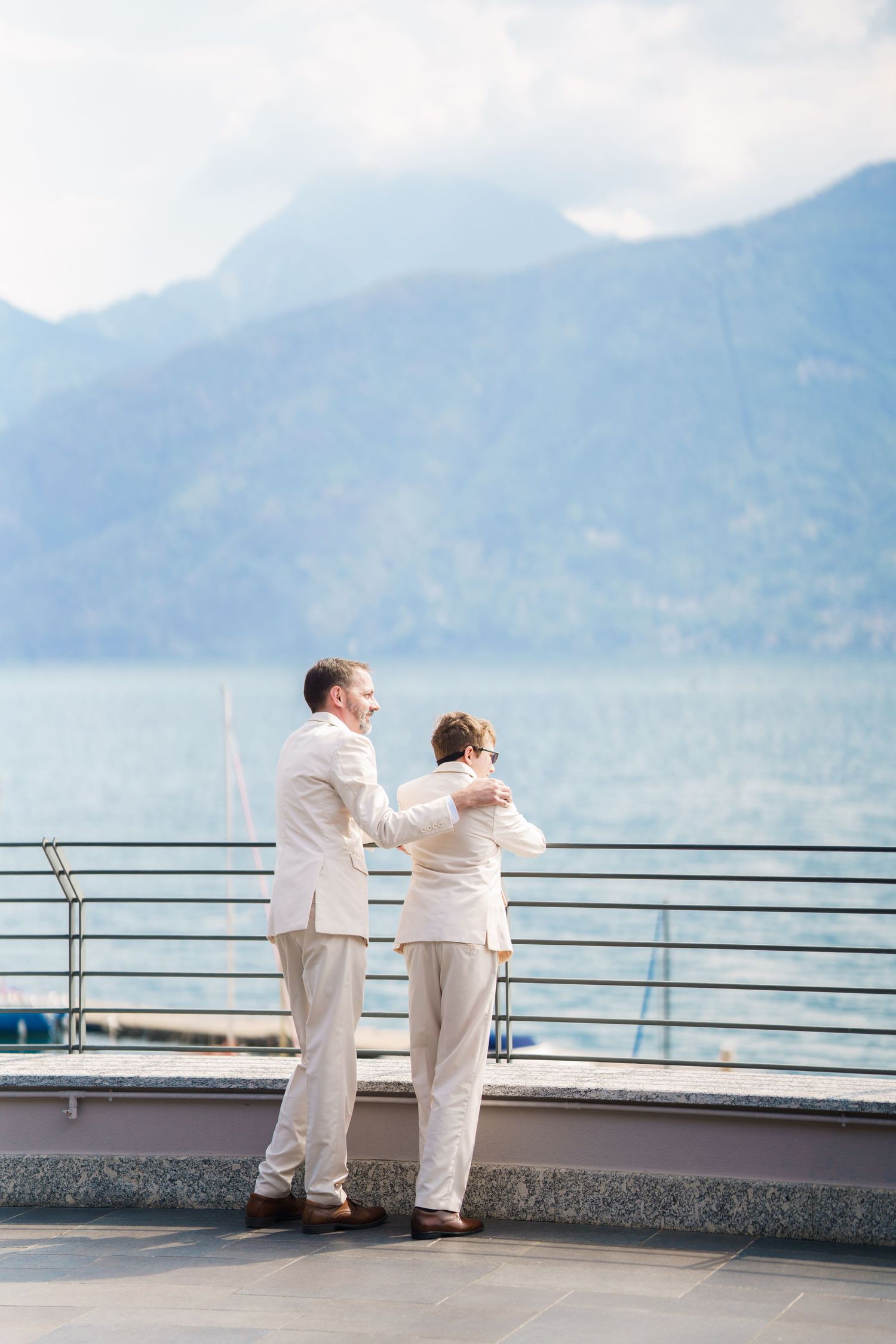 Two grooms in white suits share a tender moment on a lakeside terrace with mountain views on their wedding day.