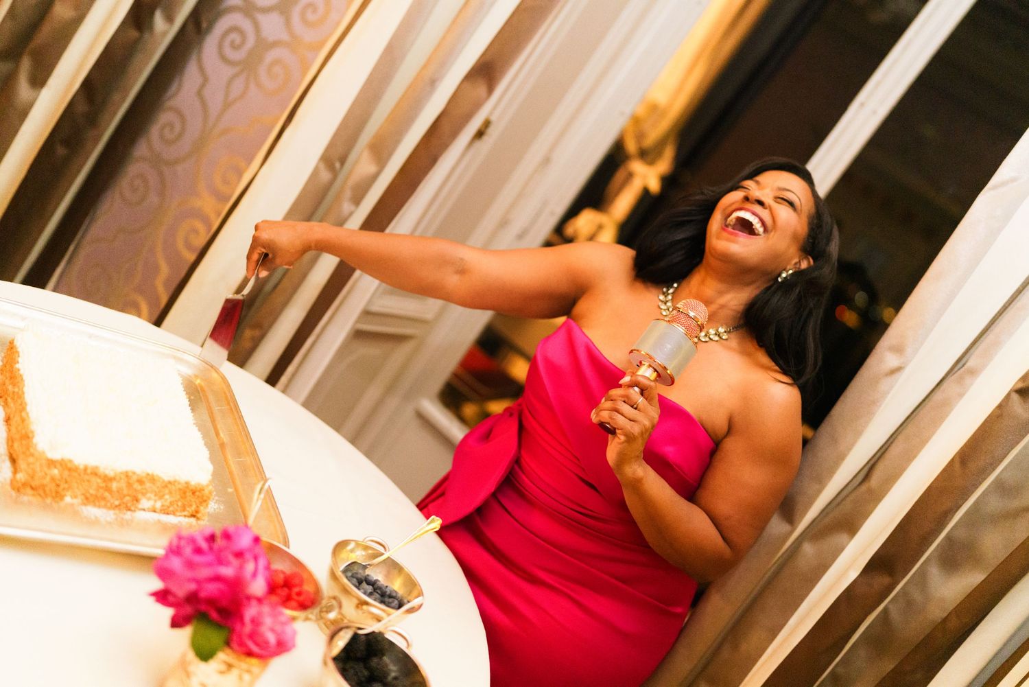 Woman in pink dress laughing joyfully at a celebration table with cake and champagne.