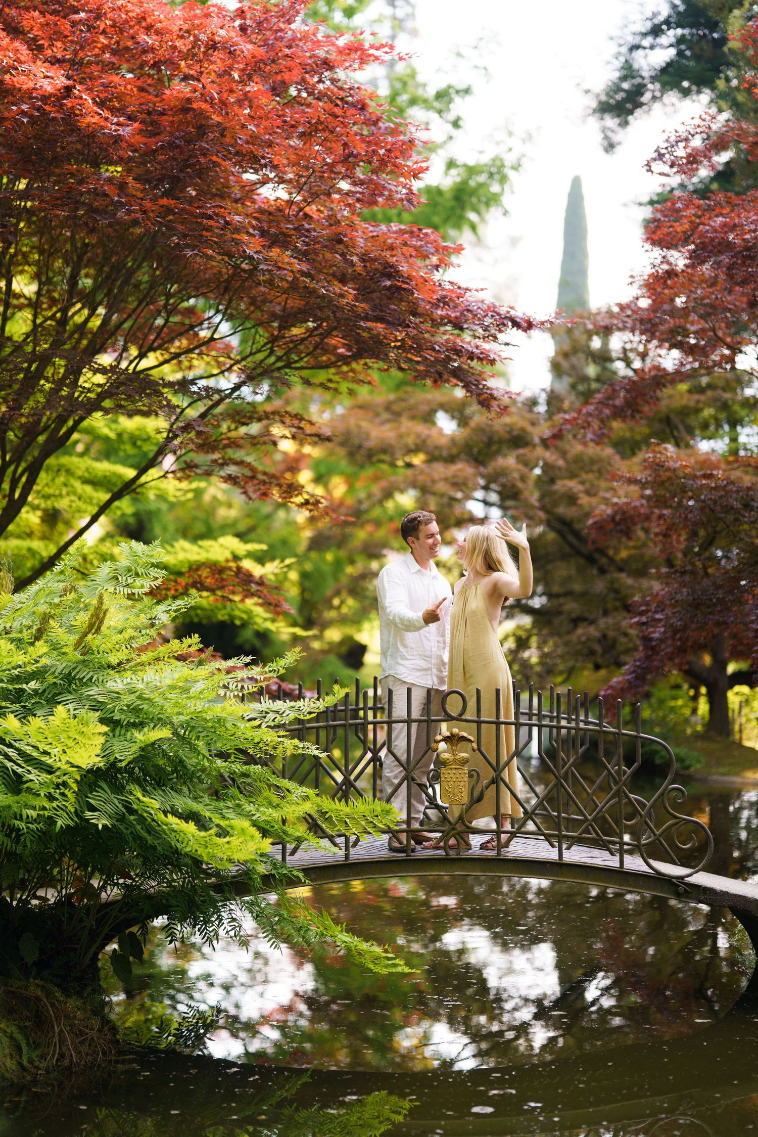 Couple standing on ornate bridge in Japanese garden with red maple trees and pond reflection.