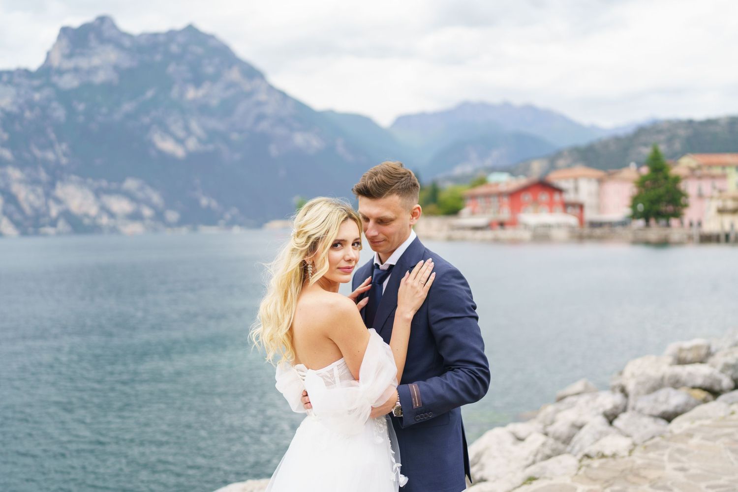 Bride and groom embracing by Lake Garda with mountain backdrop and colorful Italian village in distance.