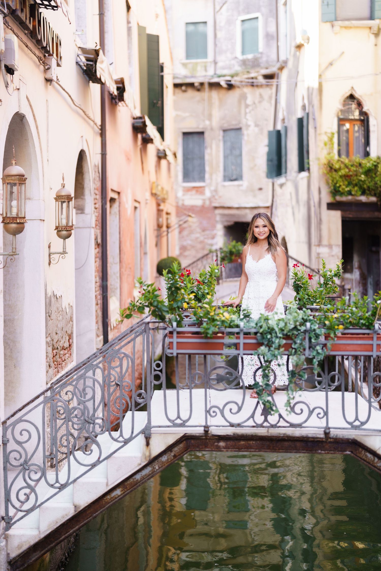 A bride in a white dress poses on an ornate bridge over a Venice canal, surrounded by colorful flowers.