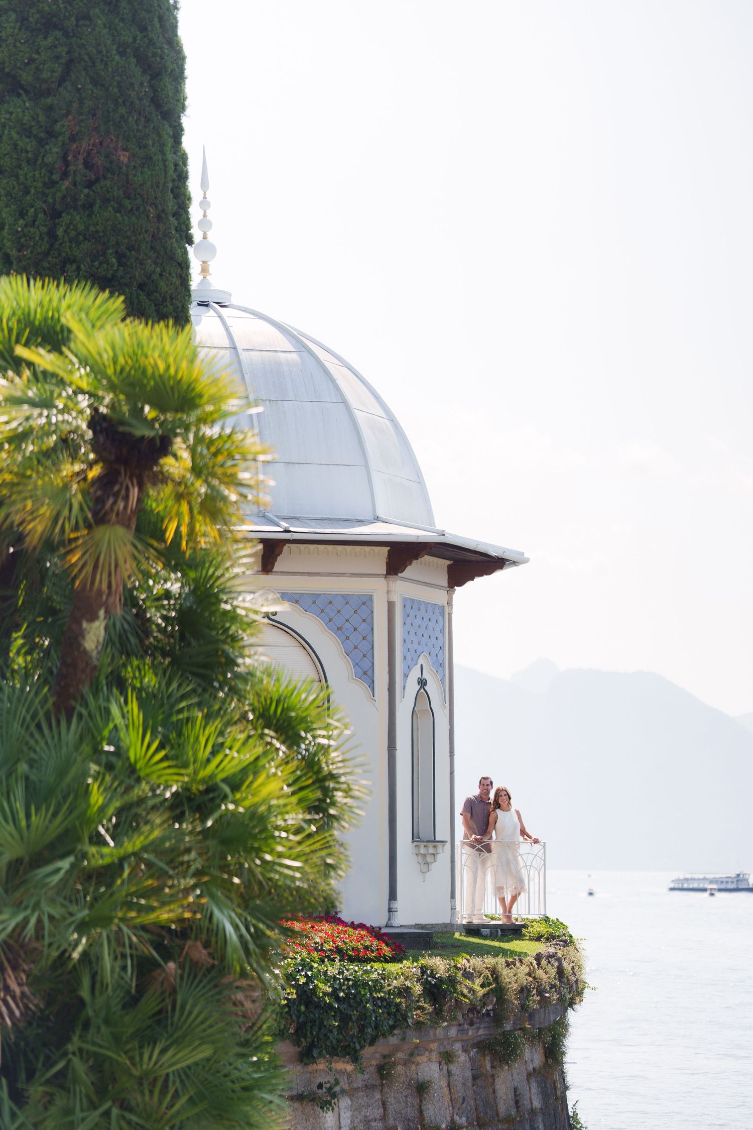Couple standing on a white domed pavilion overlooking Lake Como, surrounded by lush palm trees and mountains.