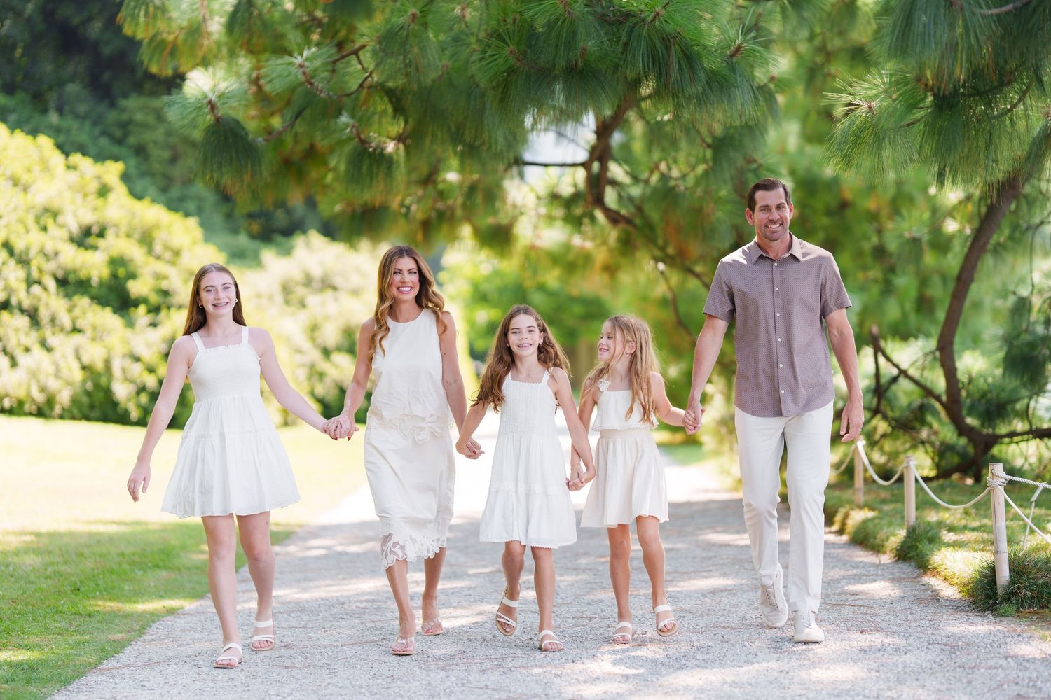 Family of five walking hand-in-hand on a garden path, wearing white outfits, surrounded by lush green trees.