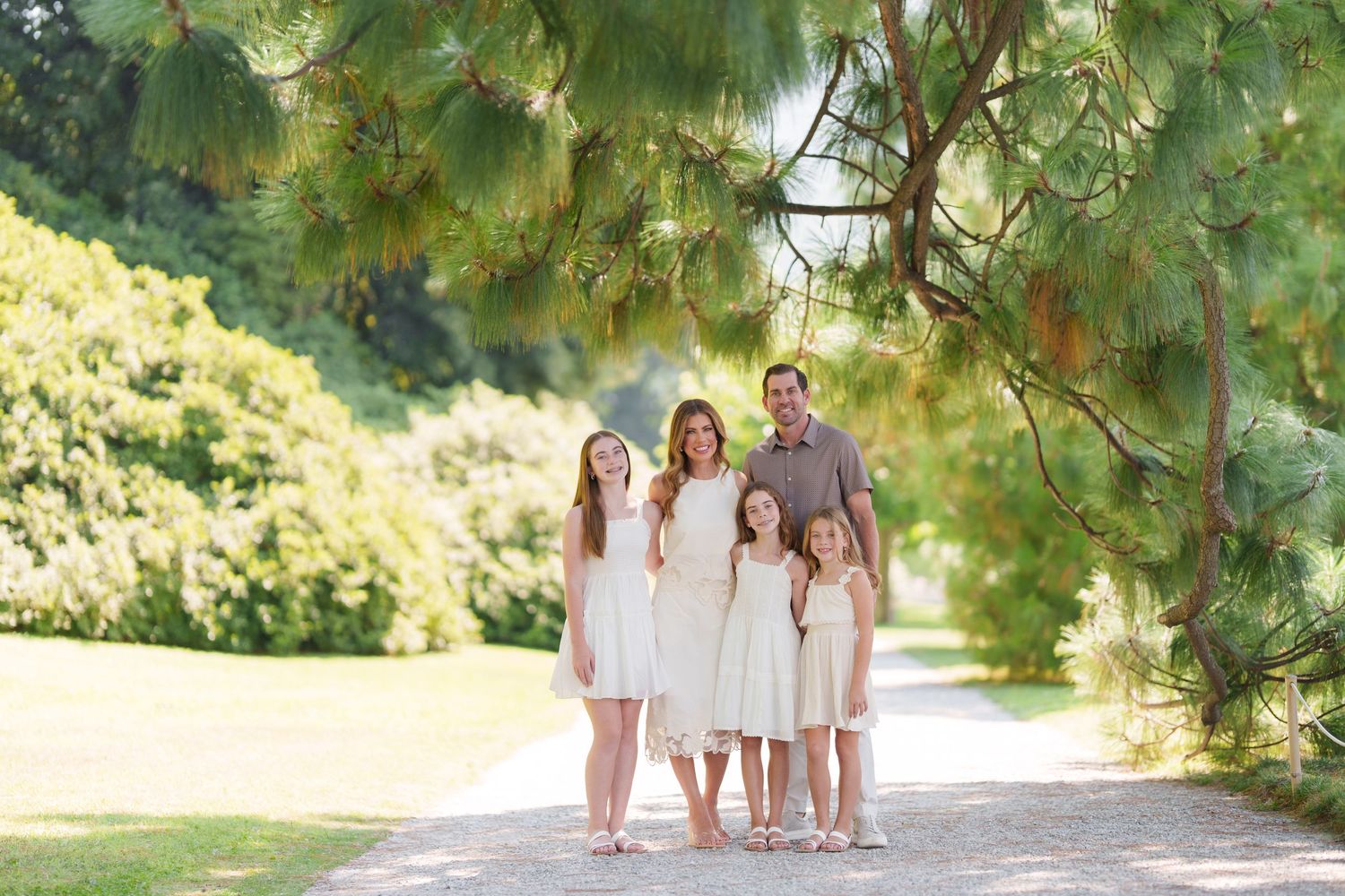 Family of five posing on a sunlit garden path surrounded by lush green trees and pine branches.