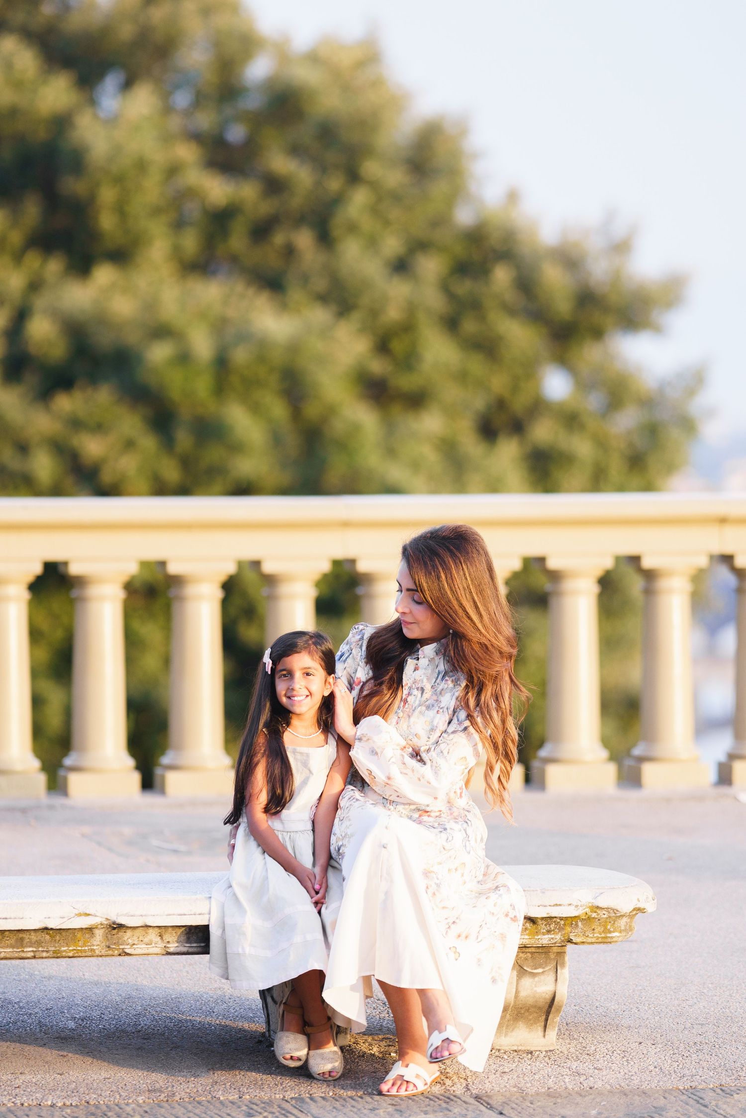 A mother and daughter in white dresses sit together on a stone bench, smiling outdoors near elegant stone columns.