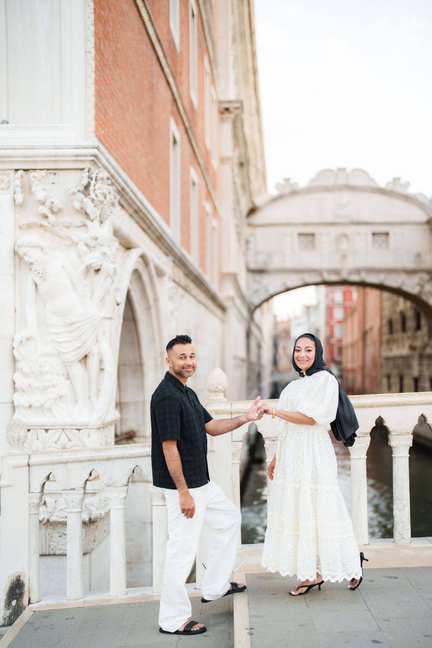 Couple holding hands near the Bridge of Sighs in Venice, woman in white dress, man in black shirt.