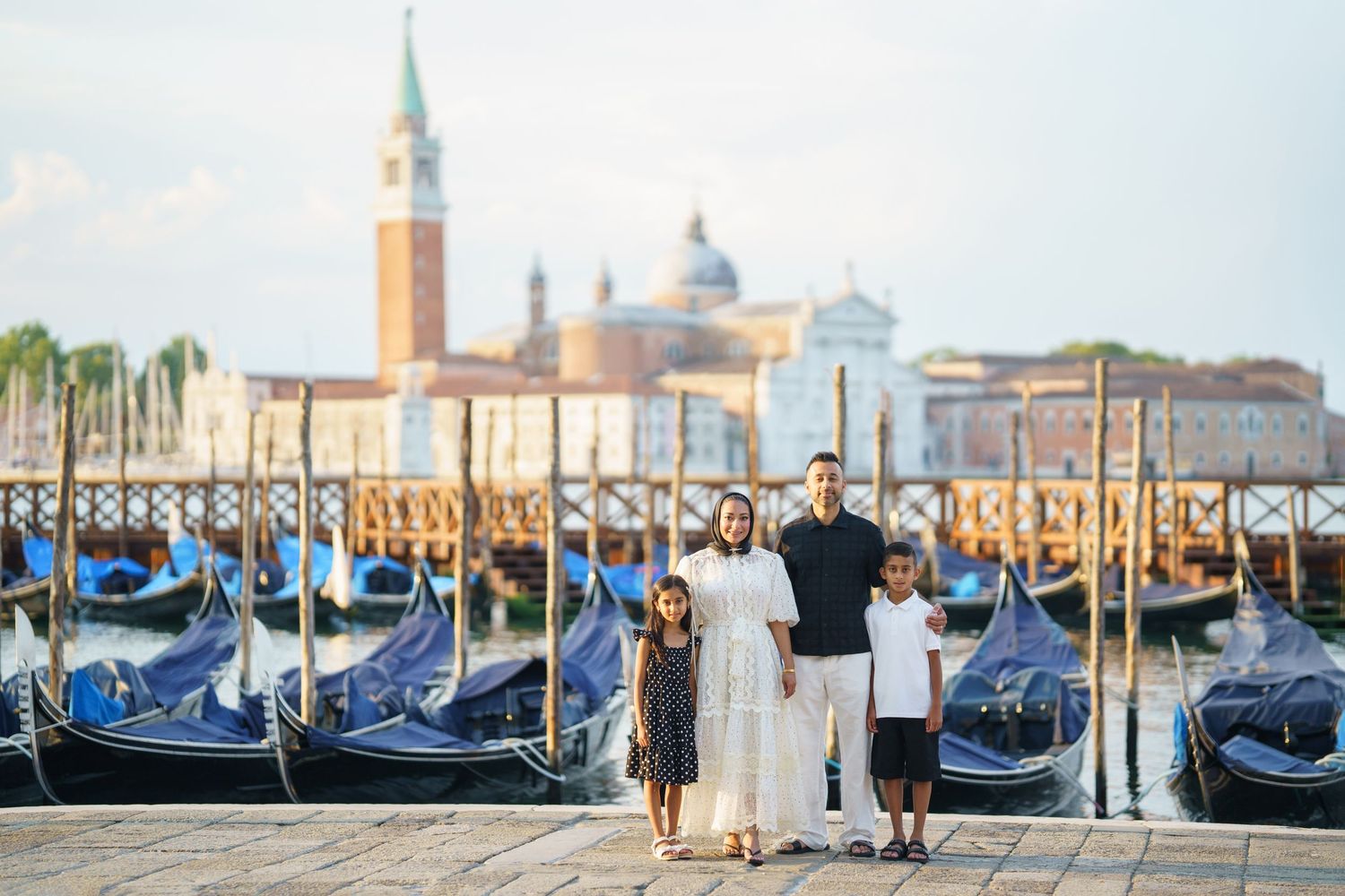Family of four posing by gondolas on Venice waterfront with San Giorgio Maggiore church in background.