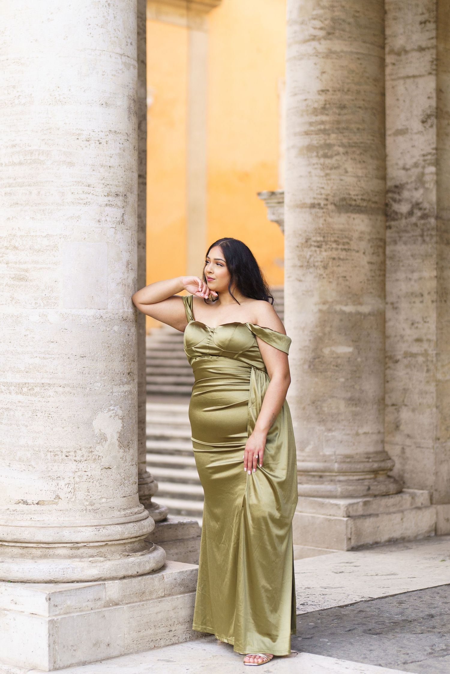 Woman in elegant olive green satin off-shoulder gown poses gracefully among classical stone columns.