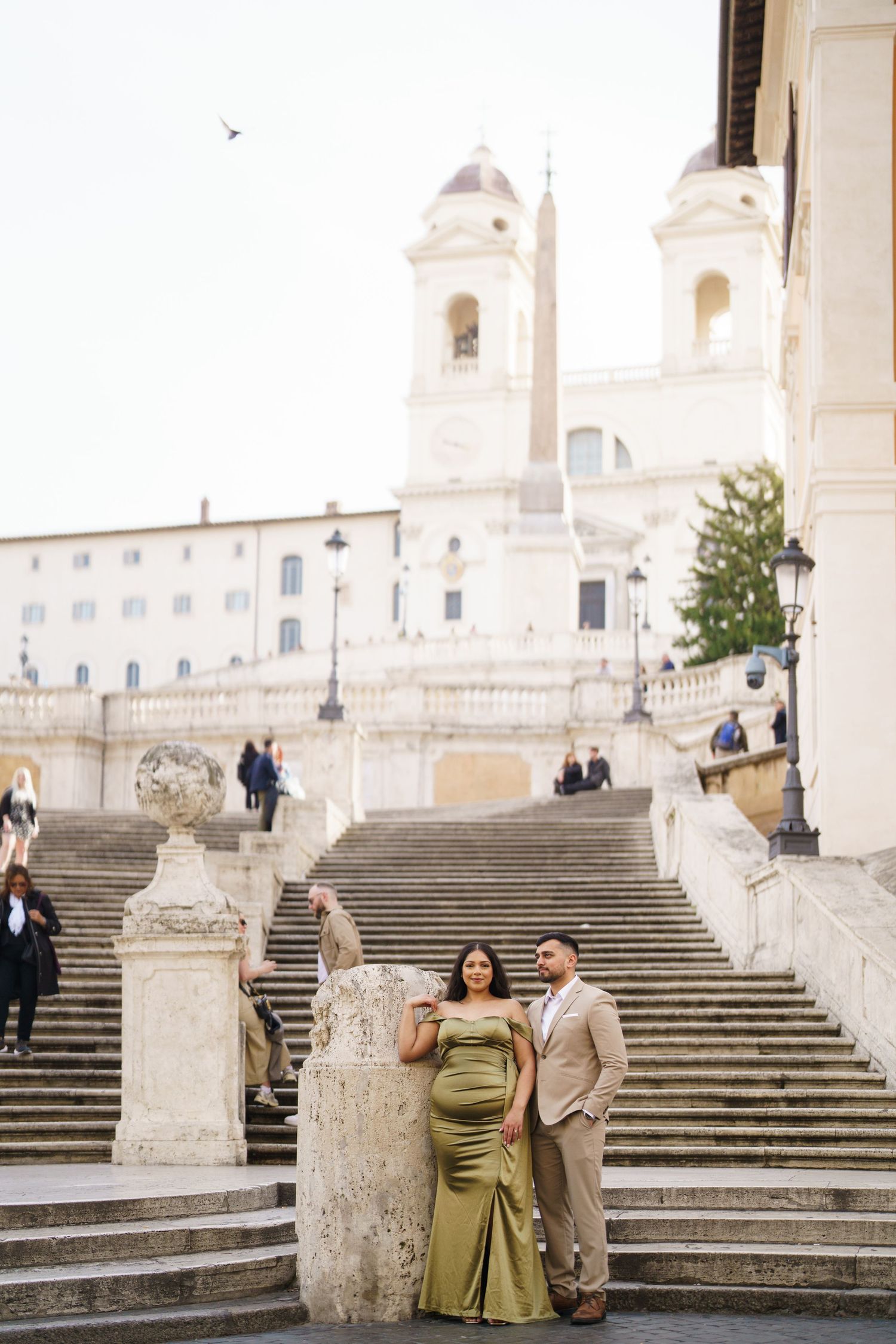 Couple posing at the Spanish Steps in Rome, woman in green gown and man in tan suit, with Trinità dei Monti church behind.