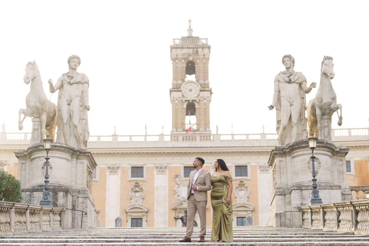 Couple embracing on steps of Piazza del Campidoglio in Rome, flanked by classical statues and bell tower.