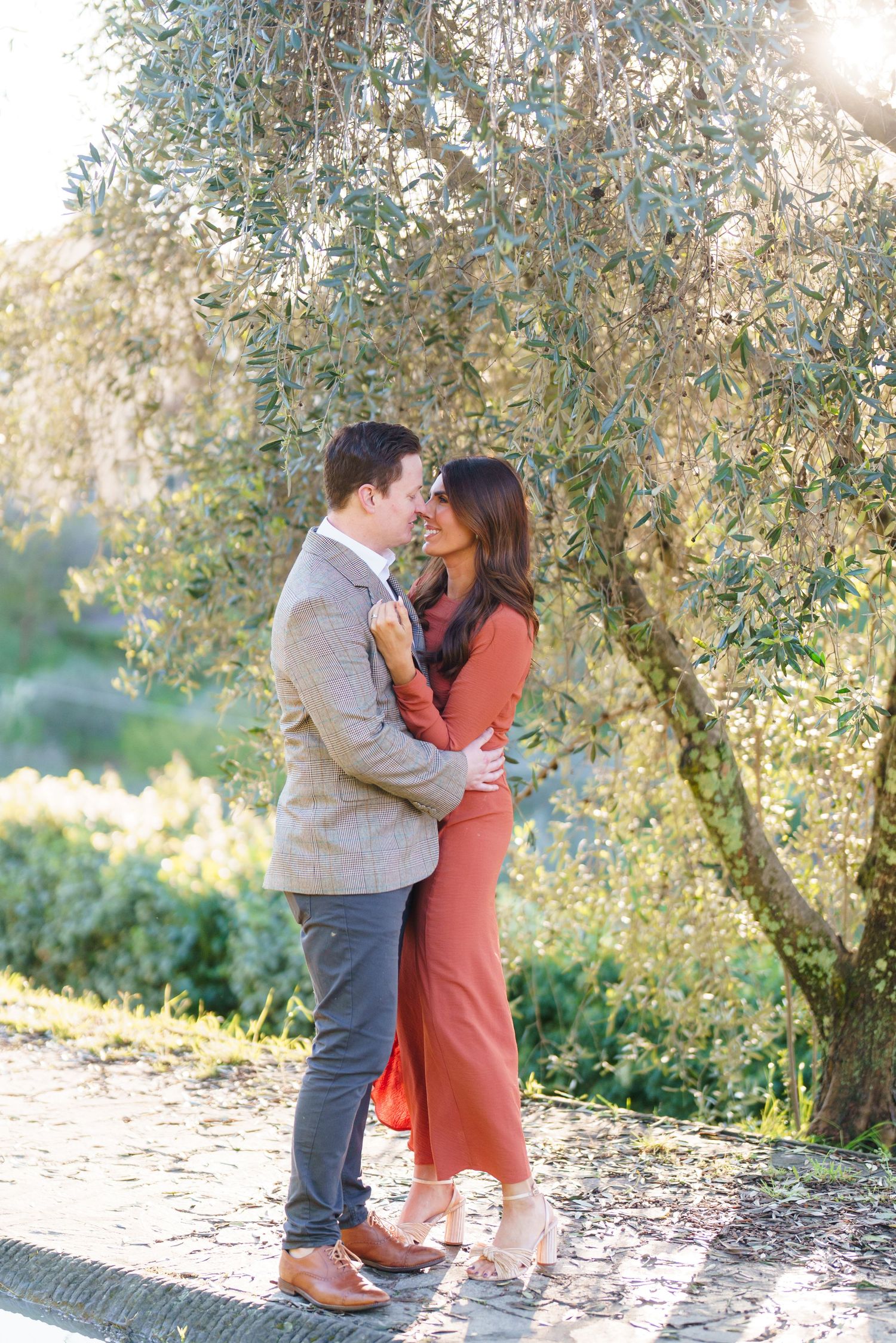 Couple embracing and gazing at each other under an olive tree, woman in rust jumpsuit, man in tweed blazer.