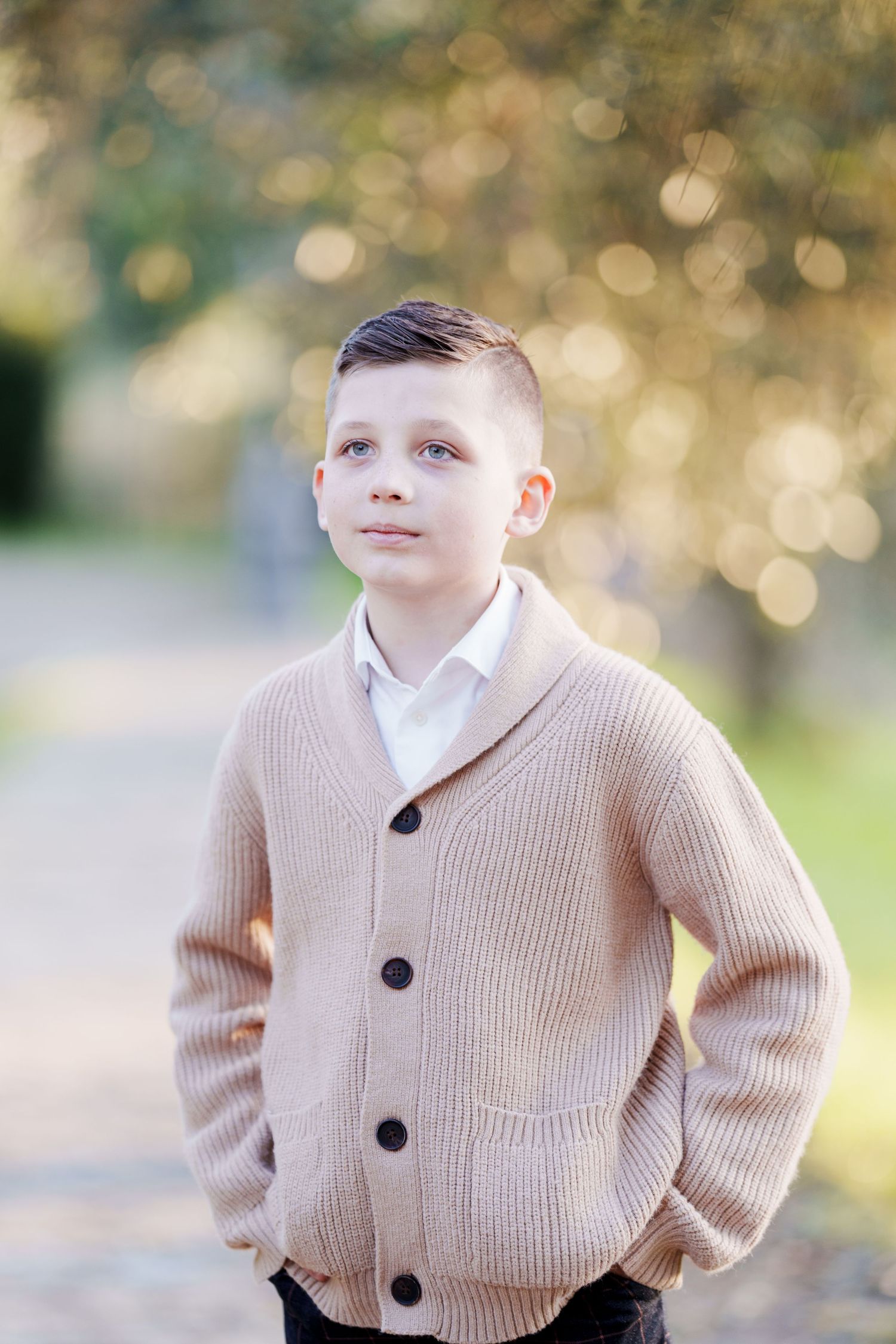 A young boy in a beige knit cardigan over a white shirt stands outdoors with hands in pockets.