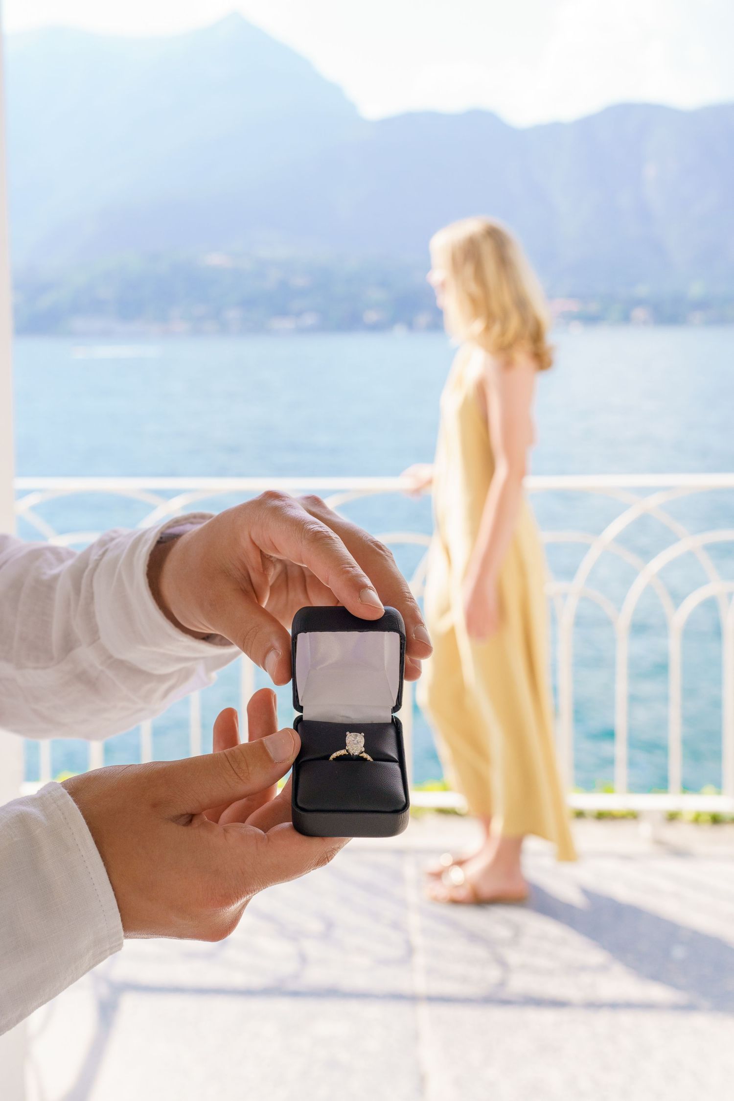 Man holding engagement ring box with woman in yellow dress standing on scenic lakeside terrace with mountains in background.