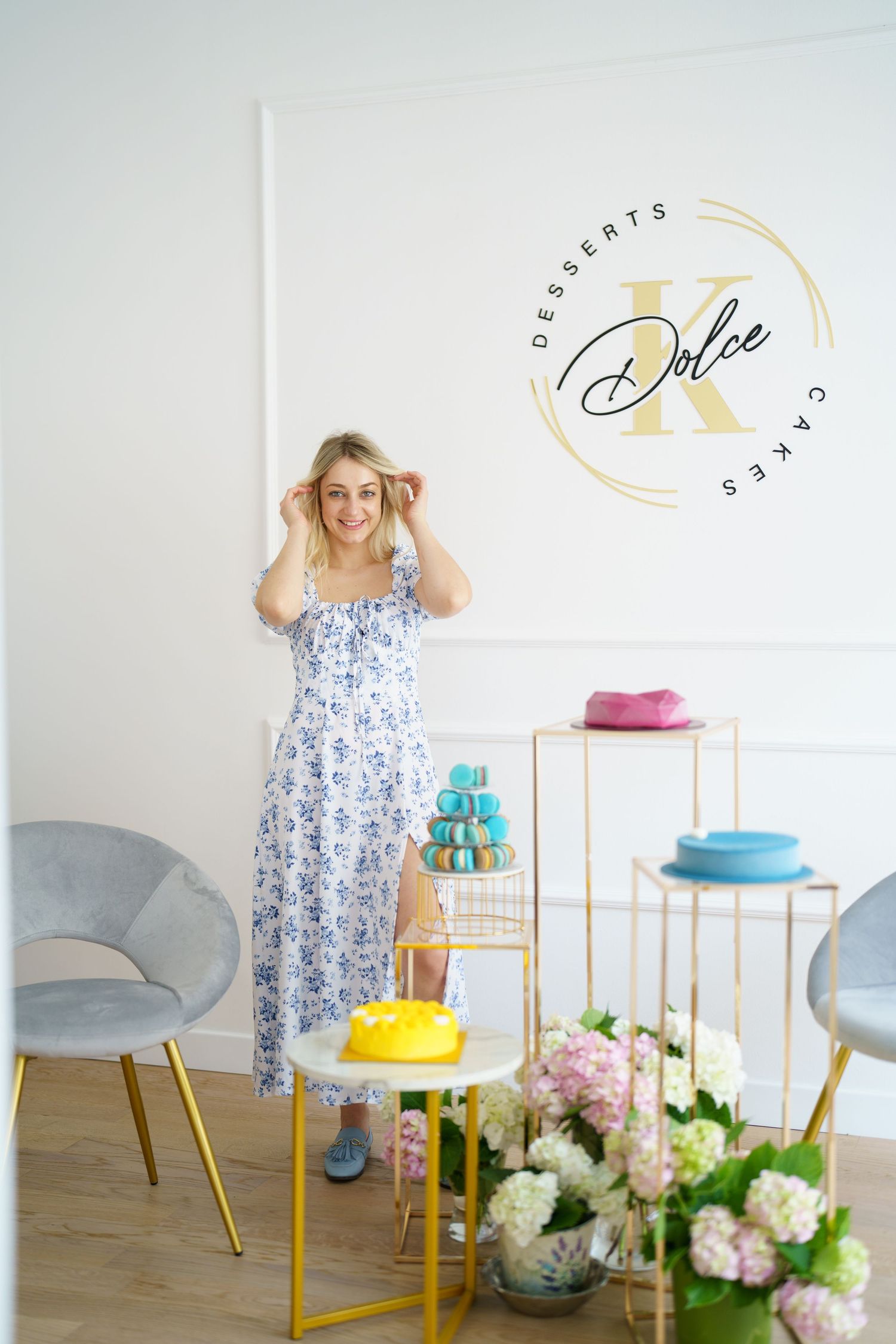 Smiling blonde woman in floral dress poses in elegant dessert shop with macarons, cakes, and hydrangeas.