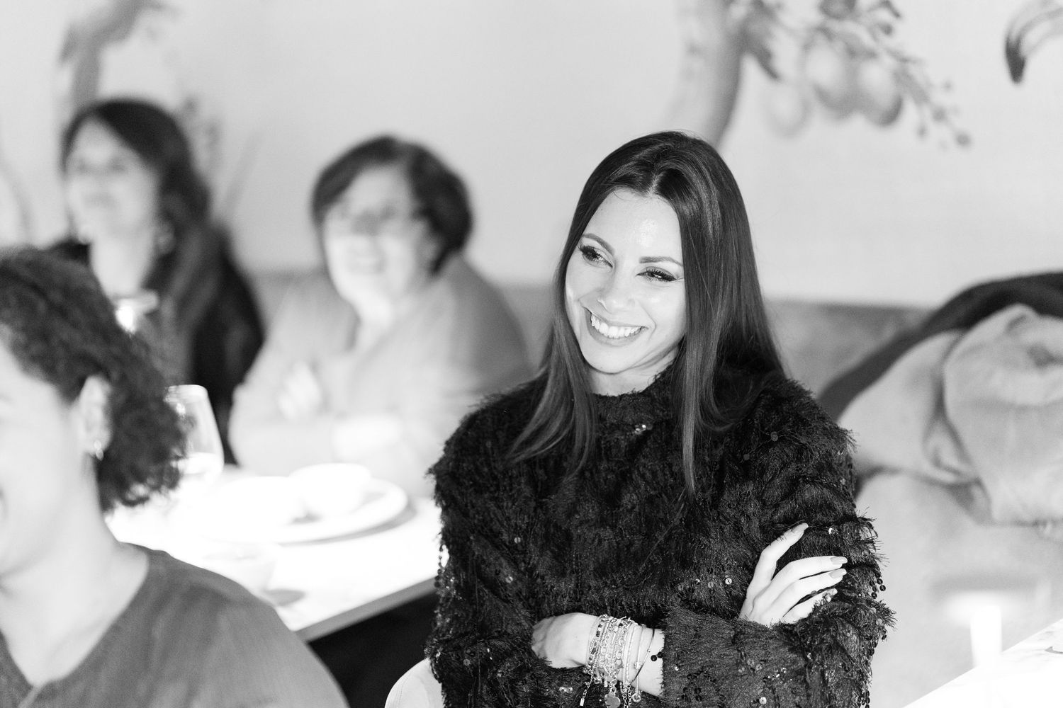 Elegant woman in black lace outfit smiling with arms crossed at social gathering.