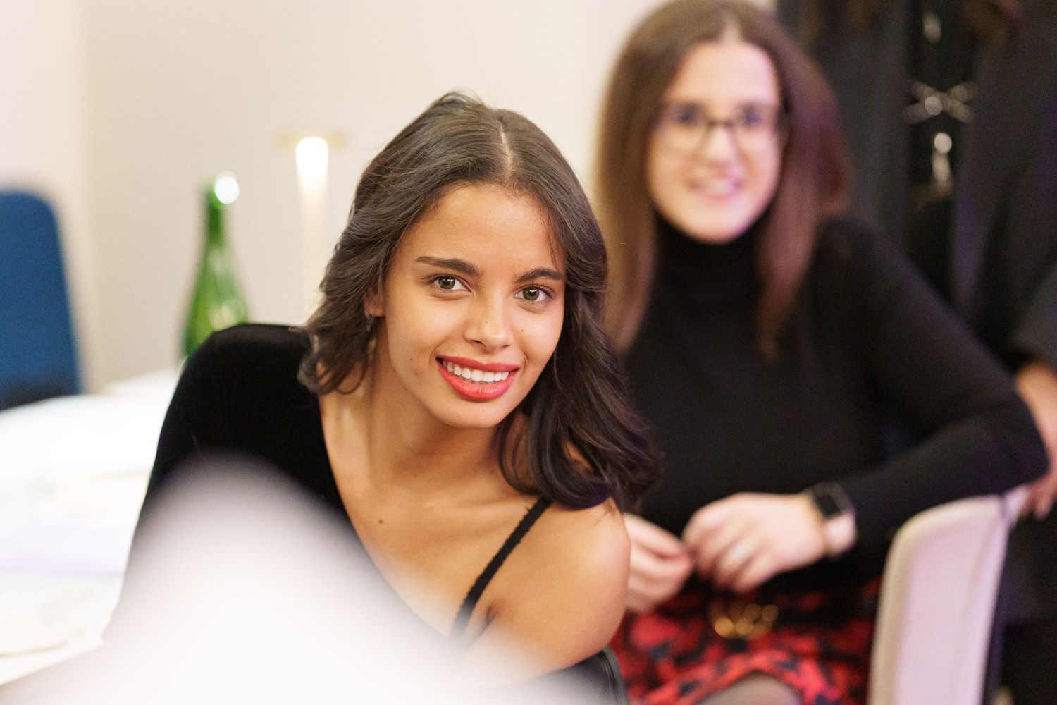 Two young women smiling at a social gathering, one in a black top with red lips, another wearing glasses behind her.