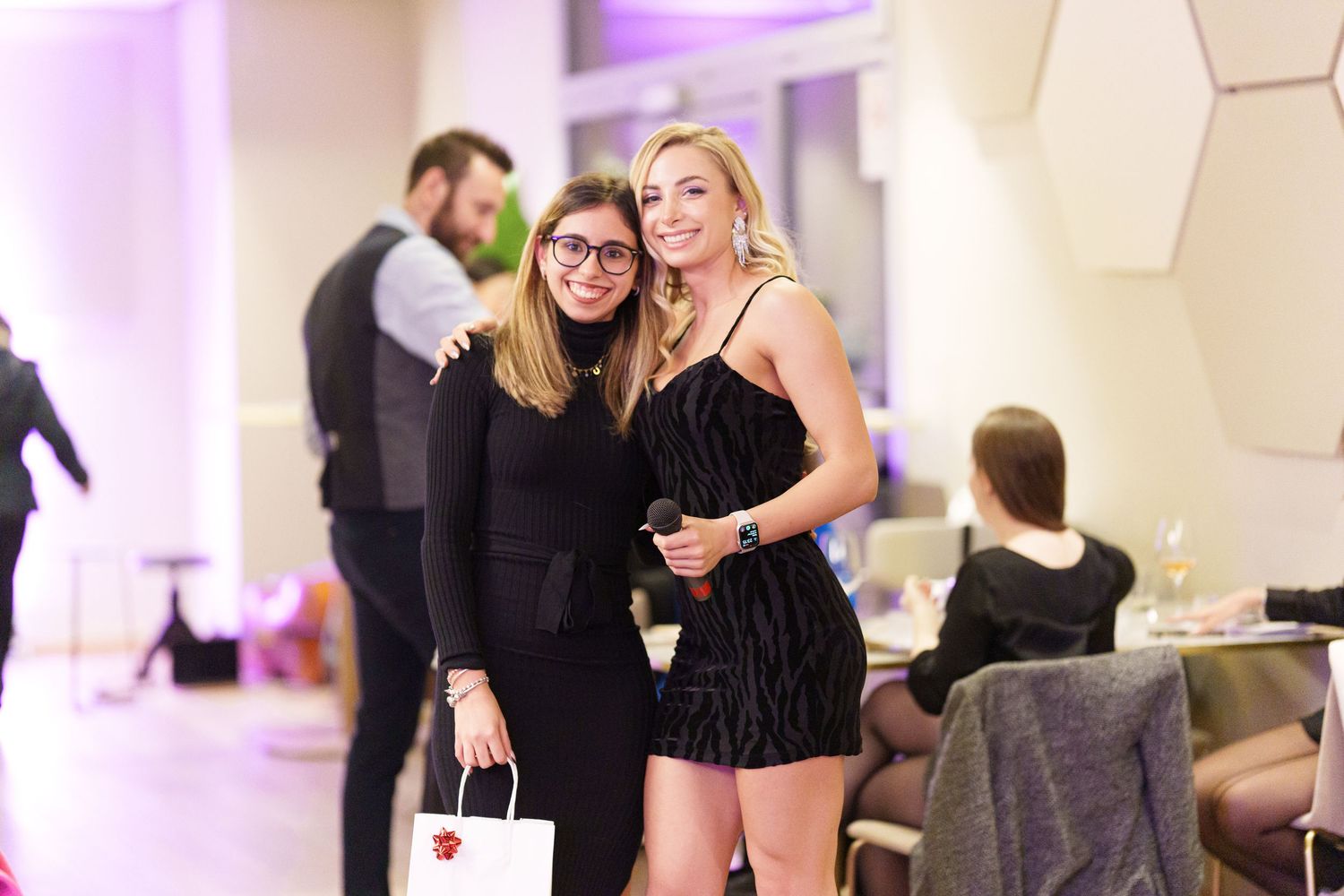 Two women in black outfits smiling together at an elegant evening event with purple ambient lighting.